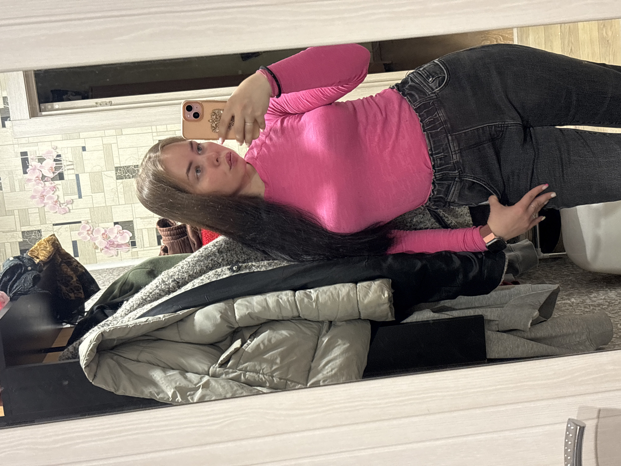 The image shows a woman taking a selfie in a mirror. She is wearing a pink long-sleeved shirt and dark jeans. She is holding a phone in her left hand. The background includes a wall with a patterned wallpaper and a pile of clothes on a shelf. No information about the location in Izhevsk, Russia is available.