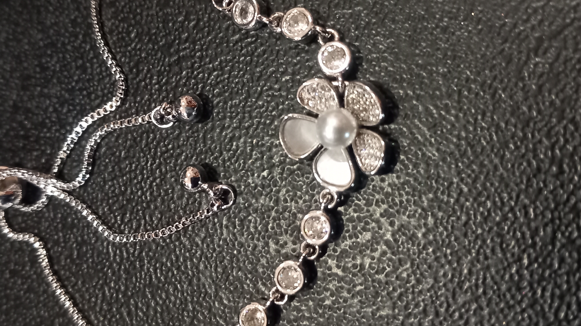 A close-up, overhead shot features a silver bracelet with a flower-shaped pendant adorned with small, clear gemstones and a central pearl. The bracelet's chain is made of small, interlinked oval shapes, with two small silver beads hanging at the end. The jewelry rests on a textured, dark surface, likely a countertop or display. The background is dark and out of focus. The scene depicts a product display, possibly in a jewelry store or market. No specific location in Kos, Greece is discernible from the image.