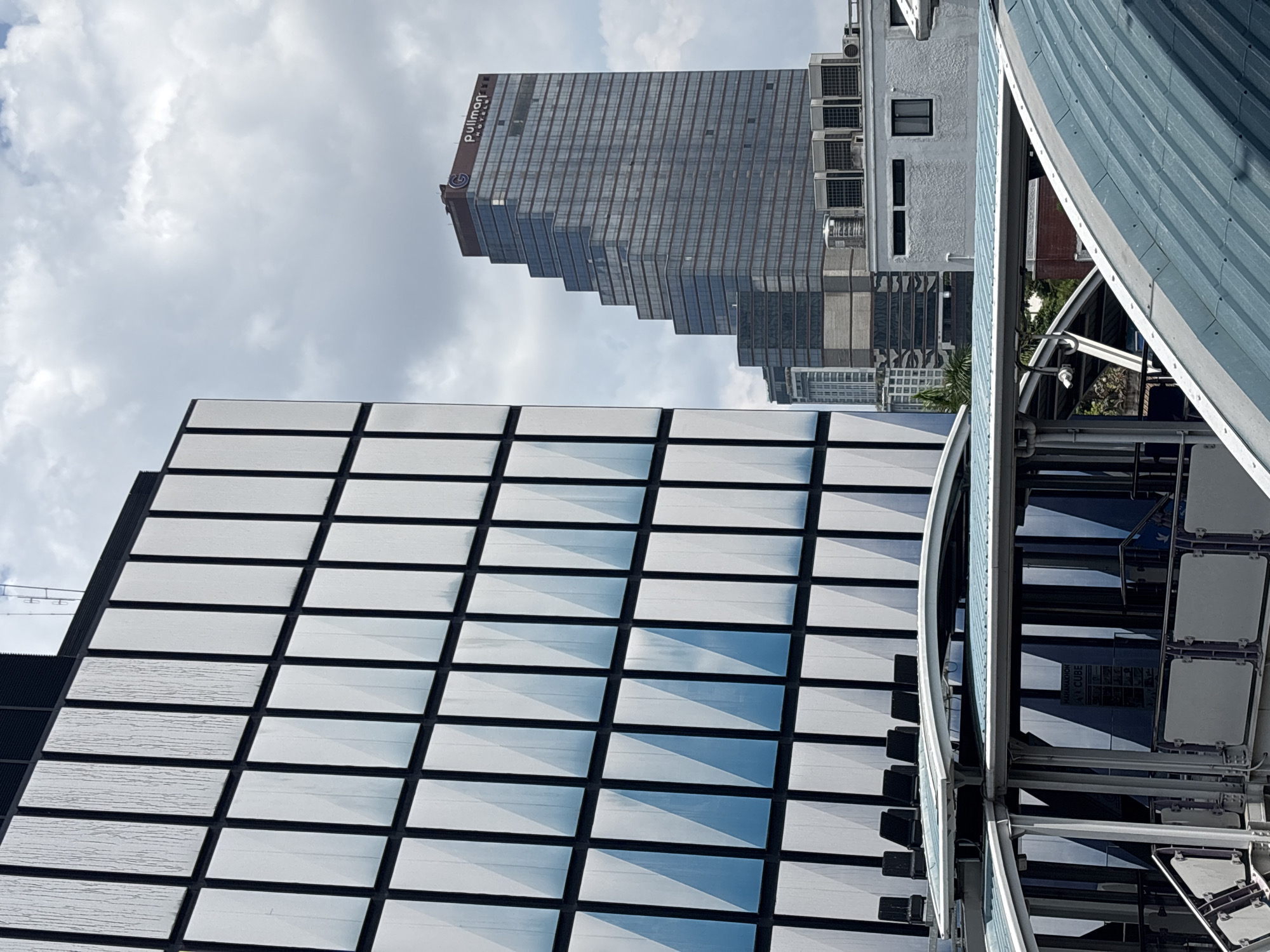 This image, taken in Bangkok, Thailand, depicts a low-angle view of several buildings against a cloudy sky. In the upper center, a tall, step-like modern glass and steel skyscraper bears the "Pullman Hotels" logo. To the left of this skyscraper, the facade of another building is visible, featuring a grid pattern with white or lightly-colored rectangular panels bordered by dark lines. A metallic structure, possibly a Ferris wheel or other construction, is positioned to the right, dominating the foreground. Its components include curved metal sheets and visible internal mechanisms. The photograph's perspective emphasizes the architecture and height of these structures against the backdrop of the sky.