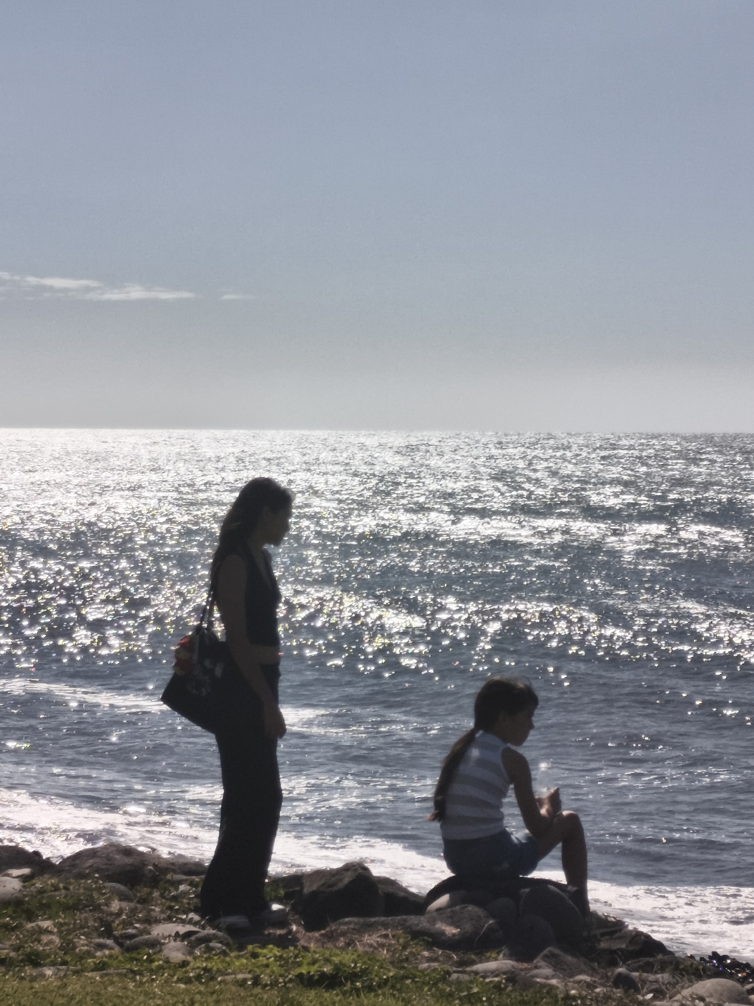 A silhouette of two individuals, an adult and a child, stand on a rocky shore overlooking a sparkling ocean. The adult, on the left, is wearing a dark sleeveless top and dark pants, with a patterned bag slung over their shoulder. The child, on the right, is seated on a rock, wearing a striped top and shorts, and appears to be holding something. The ocean surface is highly reflective due to the sun, creating a shimmering effect. The background shows a hazy sky with a faint horizontal line of clouds. The location is identified as Sainte-Marie, France, though this specific detail cannot be visually confirmed from the provided image alone.