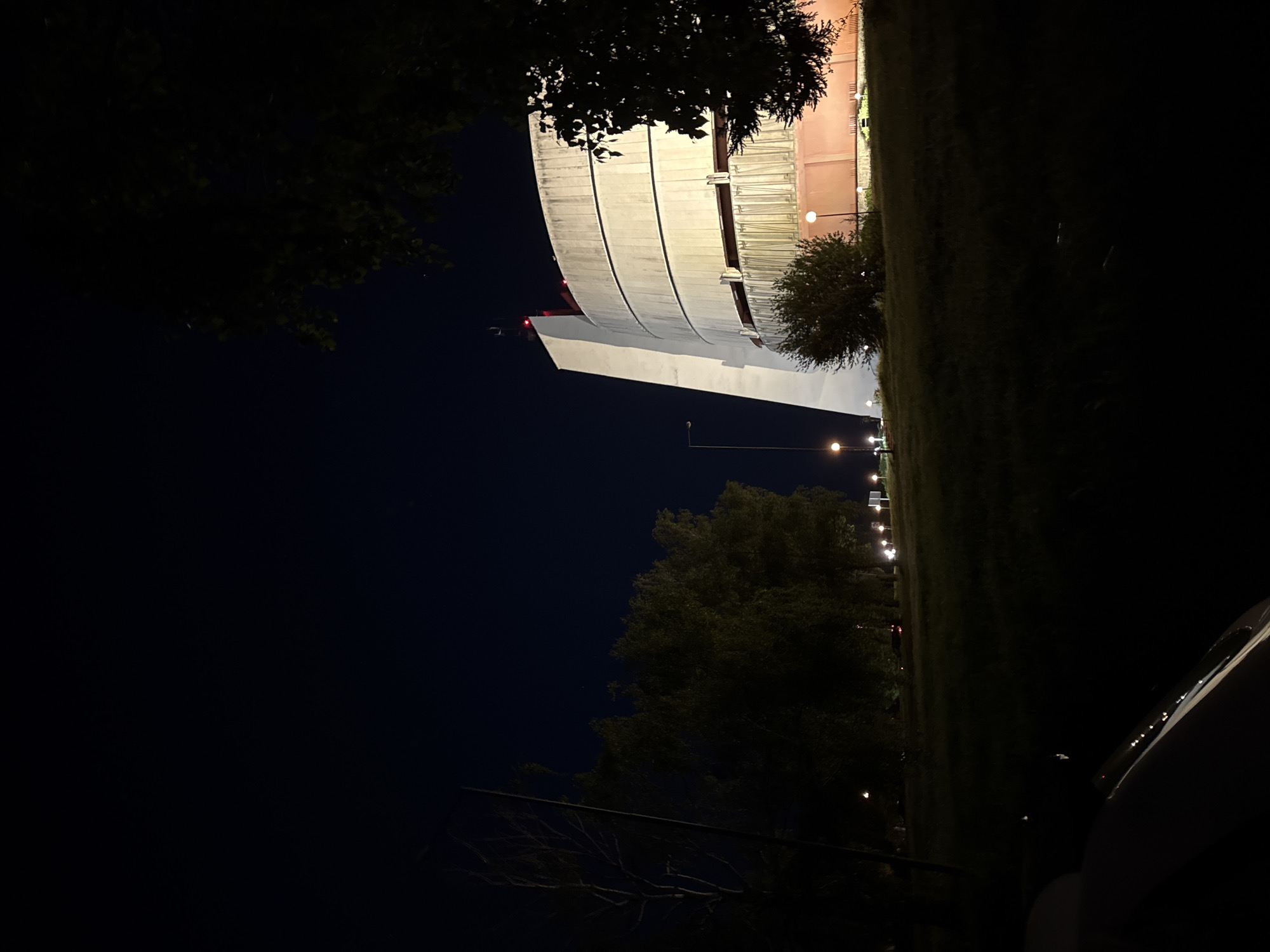 At night, a low-angle shot presents a large, cylindrical, concrete structure illuminated by artificial lighting in Pleven, Bulgaria. The structure's curved, tiered design is visible against a dark sky. The immediate foreground features silhouetted trees framing the view of the building. A grassy area to the right is dotted with evenly spaced streetlights. Part of a vehicle is visible in the lower right corner of the frame.