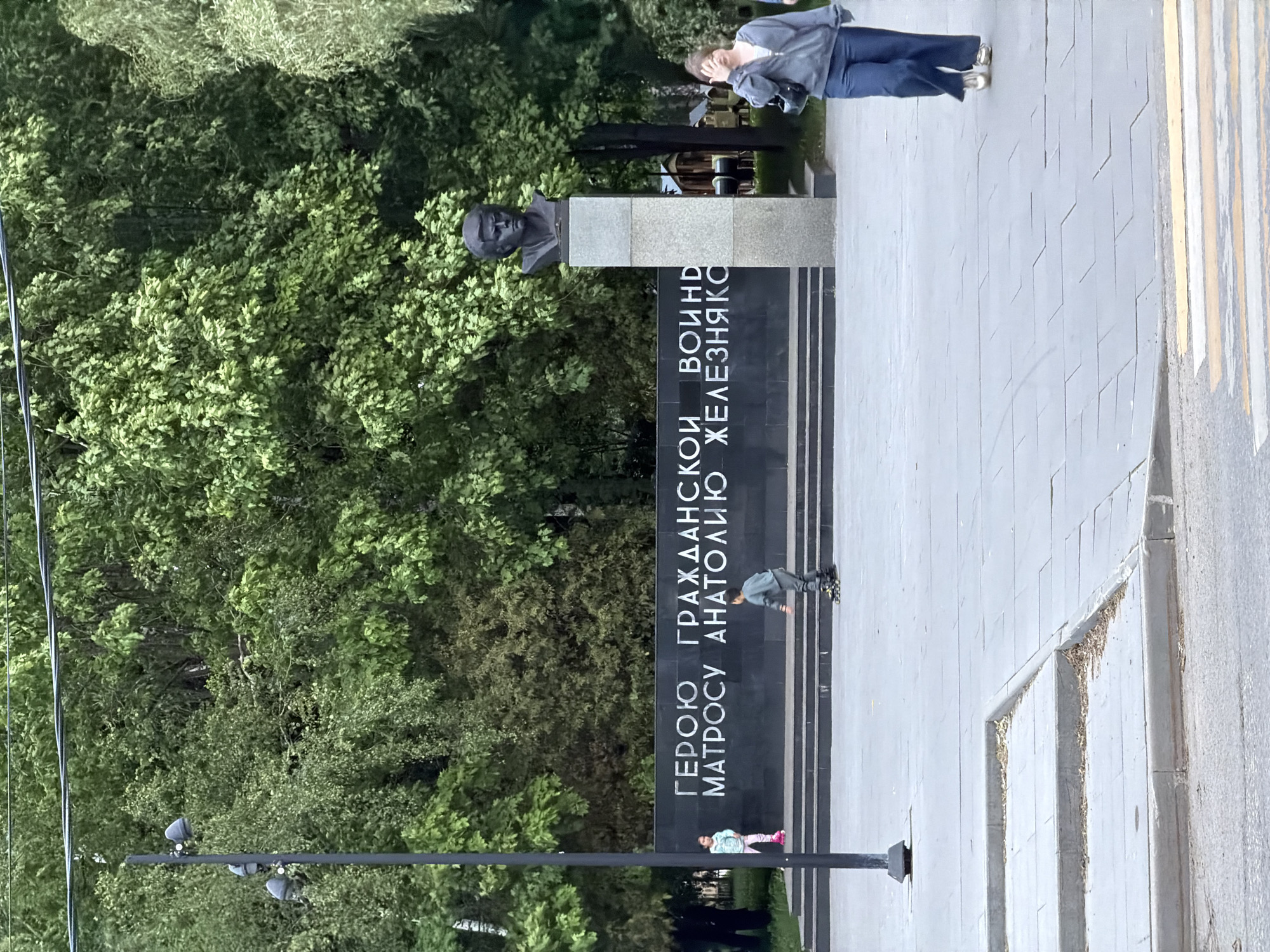 The image depicts a monument in Dolgoprudny, Russia dedicated to Anatoly Zheleznyakov, a hero of the Civil War. The monument includes a dark bust atop a stone pedestal with Cyrillic inscriptions. Several individuals are present in the scene. An adult woman stands near the bust of Anatoly. A young boy on roller skates is positioned near the monument, and a young girl is standing near the bottom of the monument. A pedestrian crossing is visible to the right. The monument is surrounded by trees.