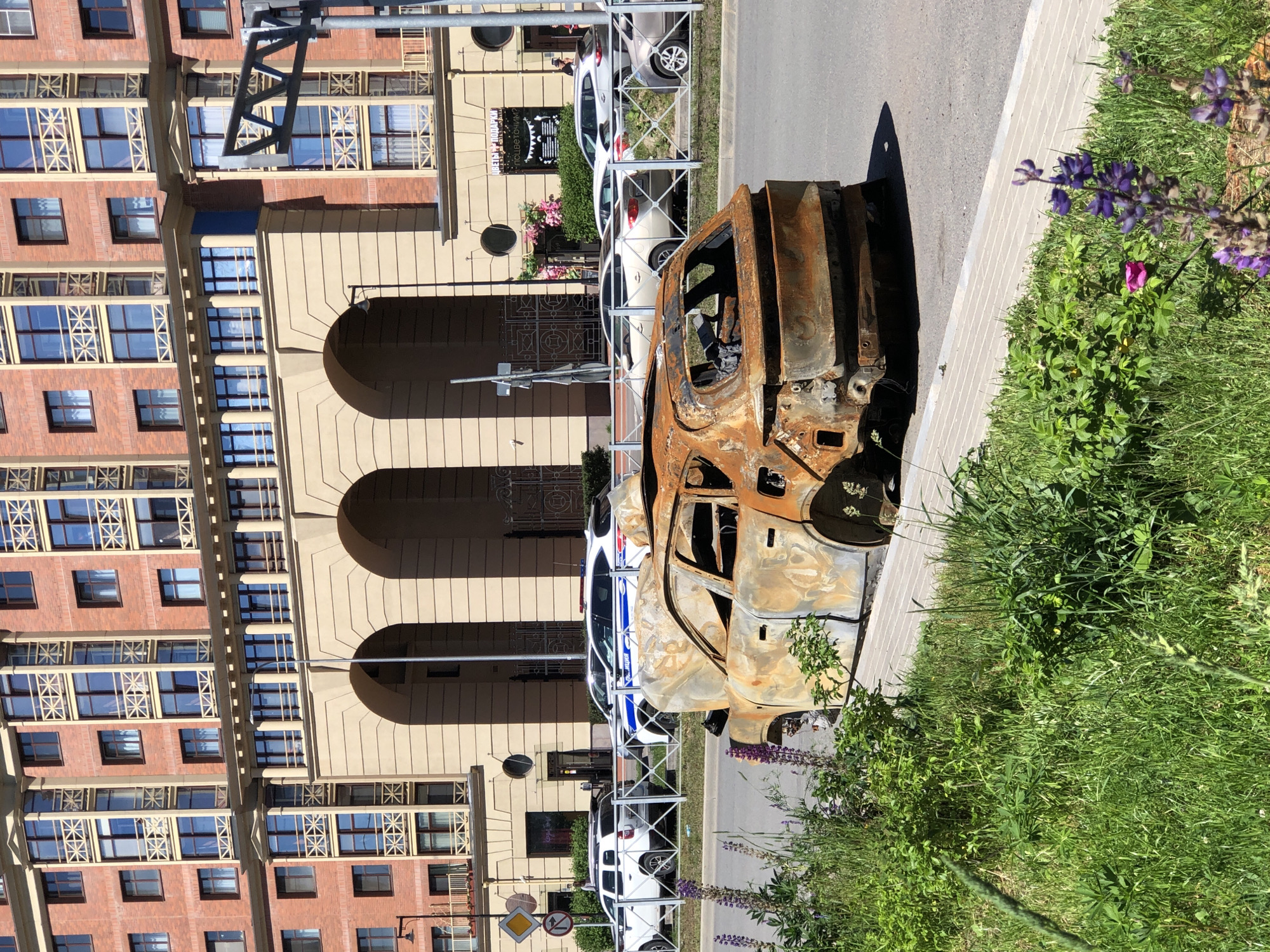 A burnt-out car sits on the side of a street in Saint Petersburg, Russia, with a large, modern brick building in the background. The car is heavily damaged, with its hood open and interior gutted, showing extensive rust and charring. A sidewalk and a grassy area with wildflowers separate the car from the street. Several other cars, including two police vehicles, are parked further down the street behind a fence. The building behind the cars features a tan, arcaded facade with multiple floors of red brick and numerous windows. The sky is clear and blue, indicating a sunny day. The scene appears to be a roadside incident or aftermath of an event.