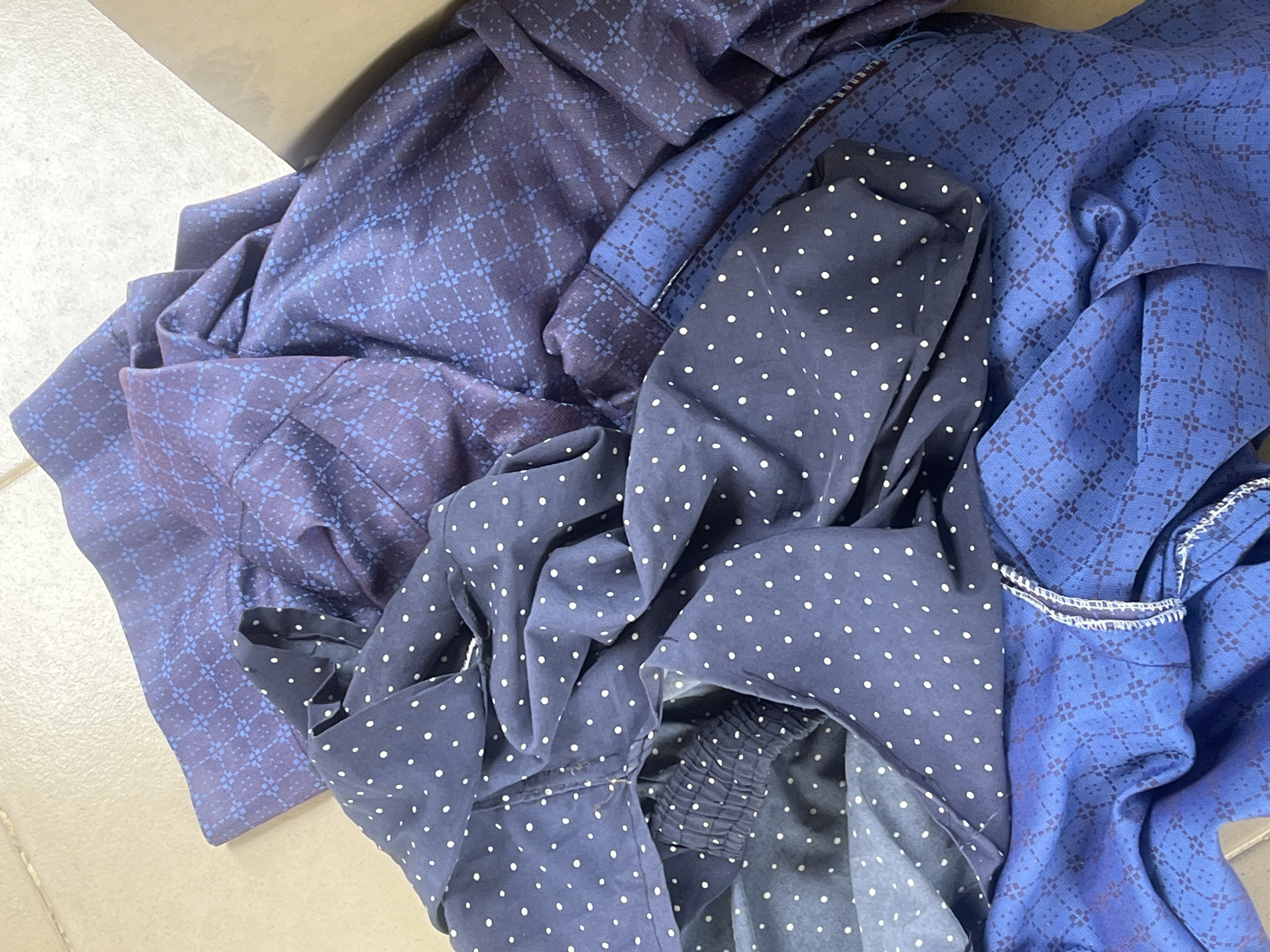 The image shows a pile of blue and purple patterned fabric, likely clothing items, folded on a tiled floor. The fabric has two distinct patterns: a dark blue with white polka dots, and a darker purple with a subtle grid-like pattern. Some of the fabric appears to be creased and rumpled, suggesting it may have been worn or is in storage. The lighting is overhead and appears to be artificial. The background shows a portion of a cardboard box. There are no people or animals visible in the image. The scene is indoors. The specific location within Jalingo, Nigeria, cannot be determined from the image alone.