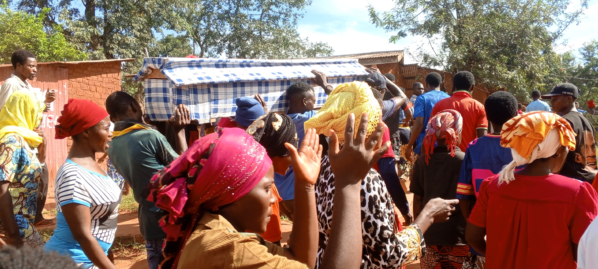 A group of people are carrying a rectangular object covered with a blue and white checkered cloth, possibly a coffin or a mattress, during what appears to be a funeral procession in a populated outdoor area. The people are of African descent and dressed in various colorful clothing, including headscarves and casual wear. Some individuals are gesturing with their hands, possibly in prayer or mourning. The environment suggests a rural or semi-rural location in the Democratic Republic of the Congo, characterized by trees, foliage, and brick structures visible in the background. The scene appears to be in Ngalula, Democratic Republic of the Congo.