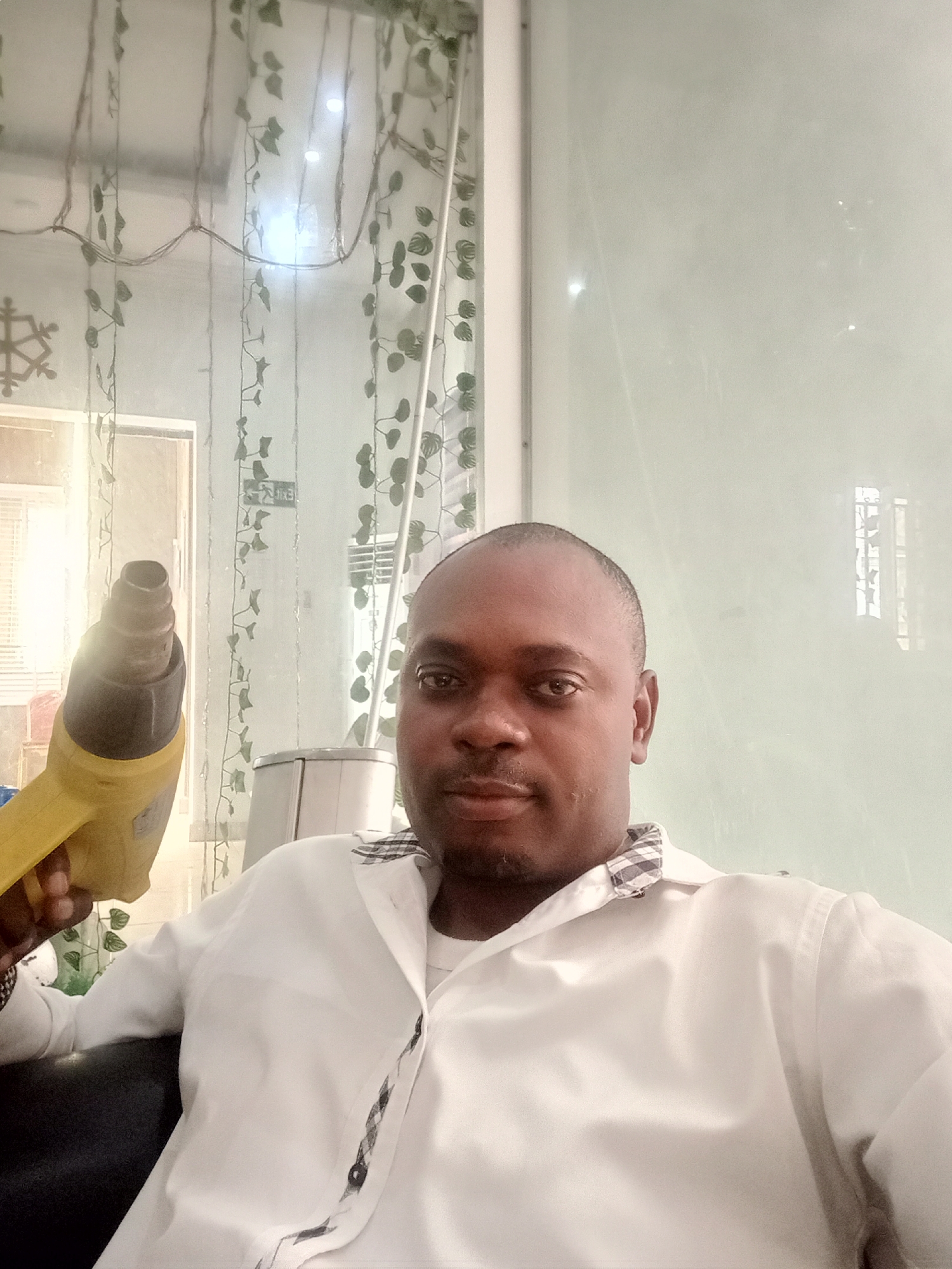 An adult male is sitting in a room holding a yellow and black heat gun. He is wearing a white shirt with a checkered pattern on the collar and placket. Behind him is a window decorated with artificial ivy and fairy lights, a door, and a frosted glass wall. The location is presumed to be in Gwarinpa, Nigeria.