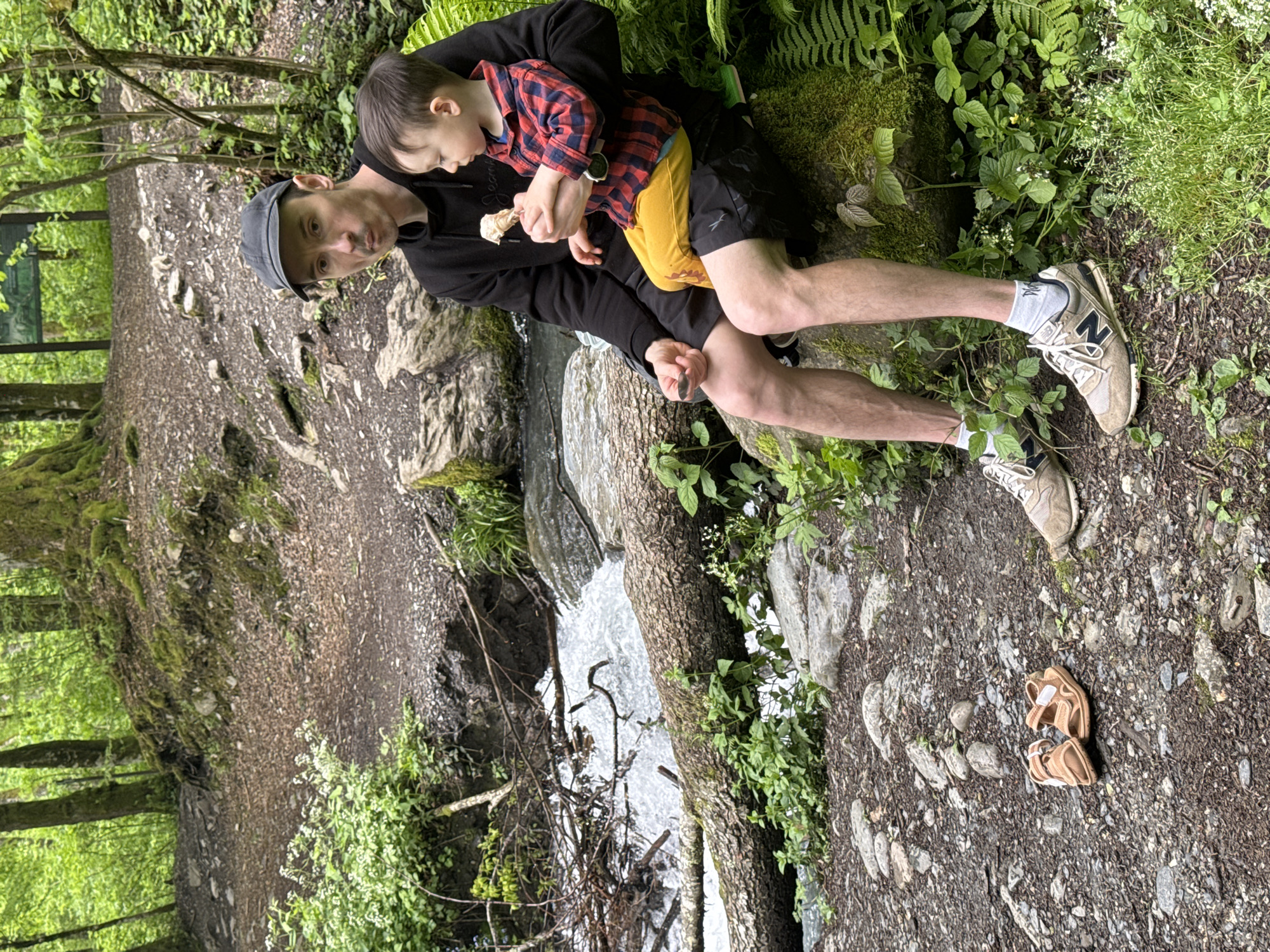 A man in a flat cap and black clothing holds a small child in a red and blue plaid shirt and yellow shorts. They are seated on a rocky ledge near a small stream. The man is looking at the camera, and the child is looking down at something in his hands. The ground around them is covered in dirt, rocks, and vegetation. A pair of sandals is visible on the ground nearby. This scene is in a wooded area, possibly in the region of Krasnaya Polyana, Russia.