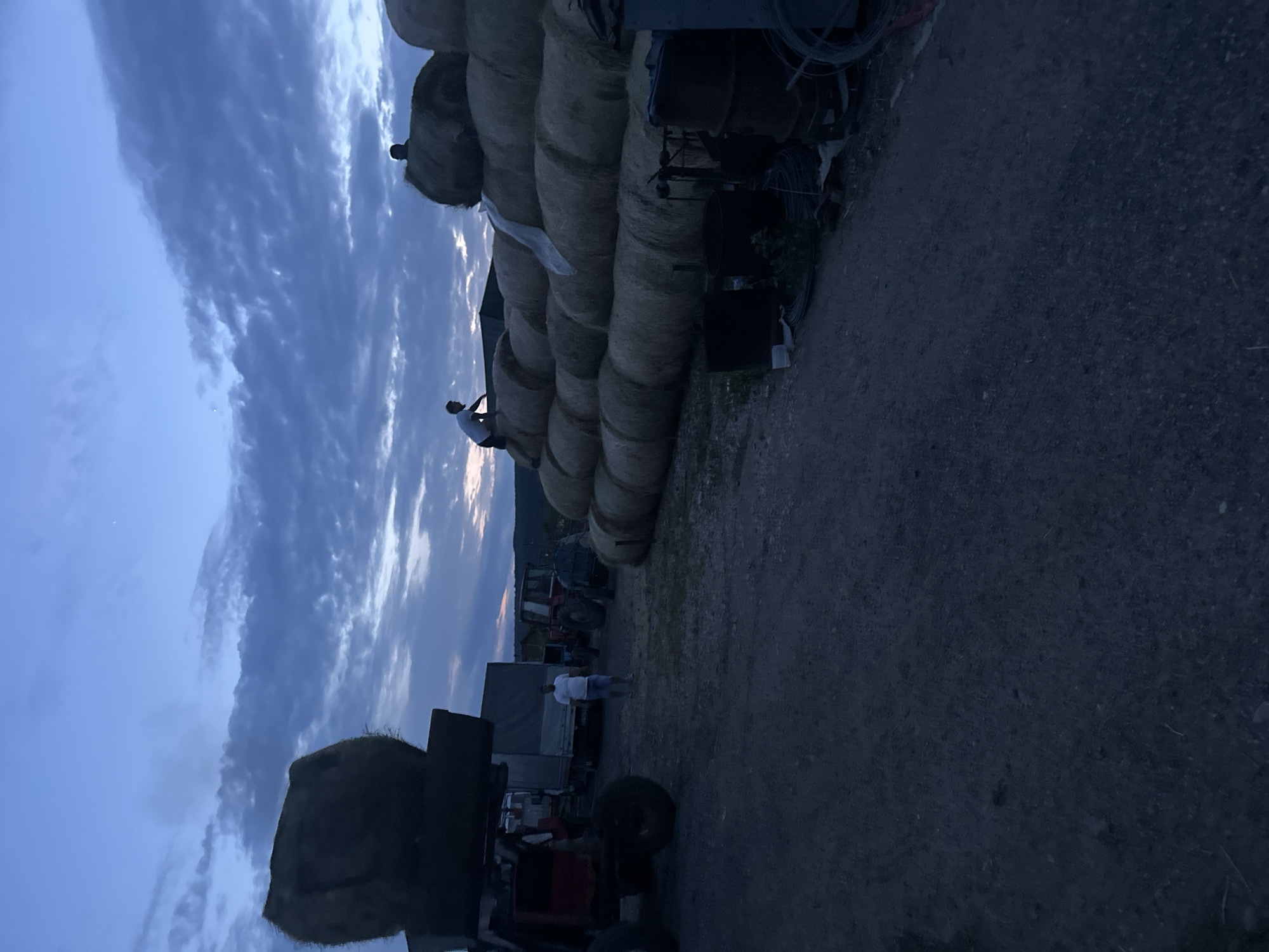 The image captures a rural, outdoor scene at dusk or twilight, likely in Chubra, Bulgaria. The sky, occupying the upper half of the frame, is a deep blue with streaky clouds and hints of lighter hues near the horizon, suggesting the last light of sunset or early dawn.
Dominating the right side of the image is a towering stack of large, round hay bales, some appearing to be wrapped in white plastic. Perched atop these bales is a silhouetted individual, seemingly observing the surroundings or taking a break. On the ground in the middle-left, another person, wearing a white t-shirt and shorts, stands near a covered truck or trailer, also silhouetted against the fading light.
Various farm vehicles and equipment are scattered across the scene. A red tractor is partially visible in the lower-right foreground. Further back on the left, other machinery, including what appears to be another tractor, and miscellaneous items like coiled wires, are present. The ground is dark, appearing to be dirt or gravel. In the far background, faint outlines of hills or mountains are discernible beneath the dramatic sky. The overall atmosphere is quiet and tranquil, characteristic of a farm or agricultural storage area at the end of the day. No visible text is present.