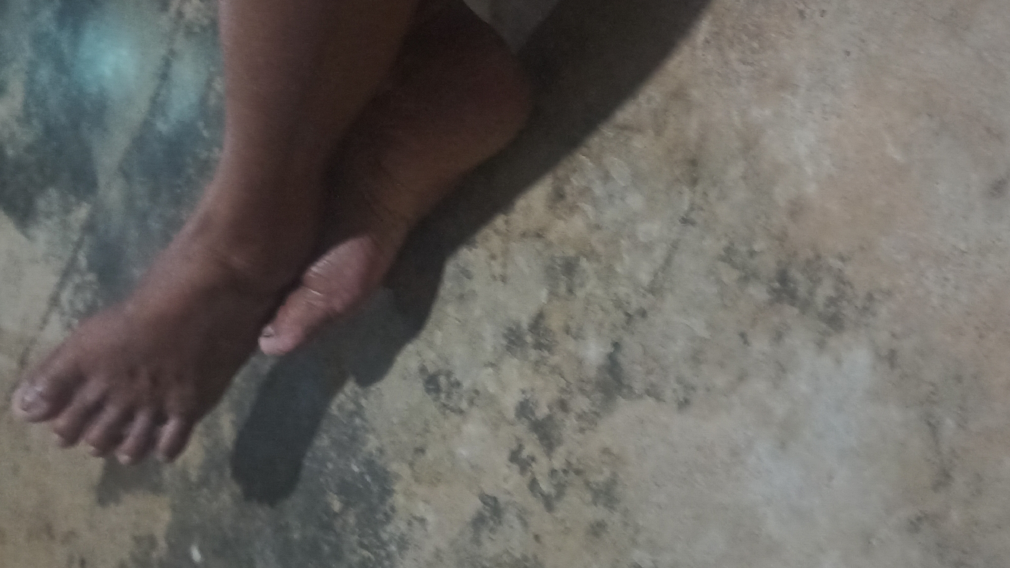 The image shows a close-up view of two feet resting on a textured, mottled floor, possibly concrete or stone. The feet appear to be positioned one over the other, with the toes of the upper foot visible and the heel of the lower foot clearly seen. Shadows are cast on the floor, indicating a light source. The lighting is dim, with some bluish light reflecting on the floor surface. The context of the location in Tuba, Indonesia, is not discernible from the visual information provided in the image.