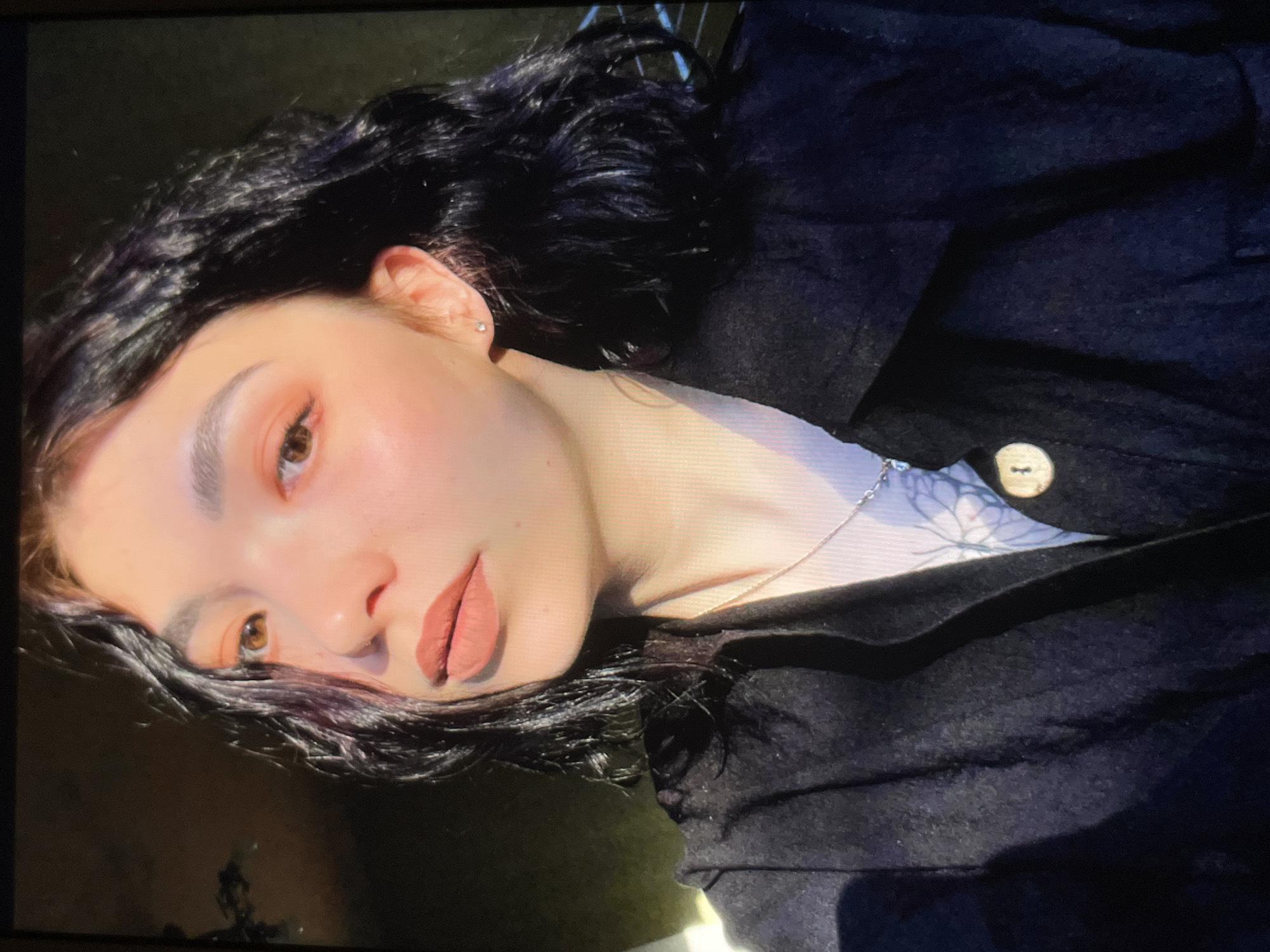A portrait of a woman with dark, wavy hair and fair skin is captured in a well-lit, indoor setting. She is wearing a dark-colored coat or jacket and a light-colored top with a floral pattern visible beneath the coat. A delicate silver chain necklace rests on her neck. The woman has light brown eyes and is wearing makeup, including peach-toned eyeshadow and nude lipstick. Her eyebrows are neatly shaped and filled in. She appears to be lying down, and the camera is positioned at an angle to her face. The location is Unknown, Unknown.