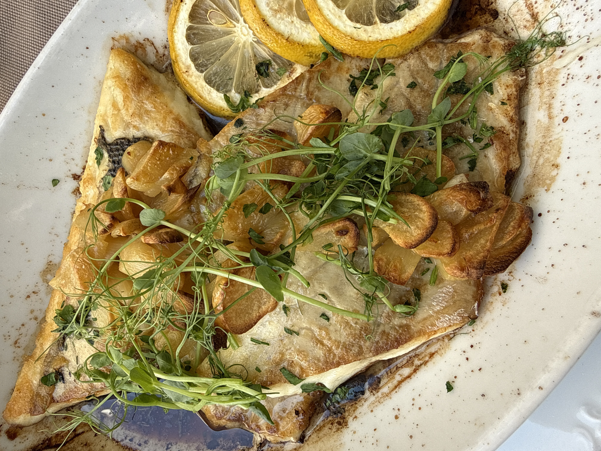 A close-up, top-down view of a prepared fish dish is presented on a rectangular, off-white ceramic plate with speckles. The dish features a large, golden-brown fillet of fish, likely baked or pan-fried, with a slightly crispy exterior. It is topped with thinly sliced, caramelized garlic and a generous amount of vibrant green microgreens. Scattered over the fish and the plate are small, finely chopped green herbs. To the right of the fish, three overlapping slices of bright yellow lemon are visible. The plate itself rests on a textured, beige surface, possibly a tablecloth. The setting suggests an outdoor dining environment, as indicated by the natural light and the presence of a woven mat beneath the plate. The overall impression is one of a beautifully presented, fresh meal. The image is believed to be taken in the town of Morinj, Montenegro.