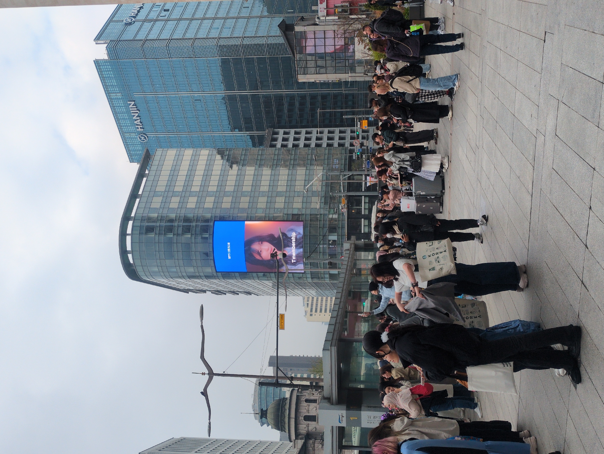 Here is a detailed description of the image:
**Environment:** The photo appears to be taken in Seoul, South Korea, as evidenced by the "HANJIN" sign on a modern glass skyscraper and the "KOREA" branded shopping bags visible in the crowd. The weather looks overcast with a gray sky.
**People:** A long line of individuals is waiting on a paved area resembling a sidewalk or plaza. The people are diverse in age, clothing, and appearance. Many individuals are carrying luggage, shopping bags, or backpacks, suggesting they may be tourists or travelers.
**Objects:** There are several tall, modern buildings in the background, indicative of an urban setting. A large electronic billboard displays a person's image with Korean writing. Streetlights and some greenery are visible.
**Actions/Interactions:** The main action is that people are waiting in line. They seem patient, and some are using their phones, suggesting they are passing time while waiting.
**Overall Impression:** The image portrays a scene from a busy urban area in Seoul. The long line of people suggests they may be waiting for something of interest, possibly a popular event or attraction.