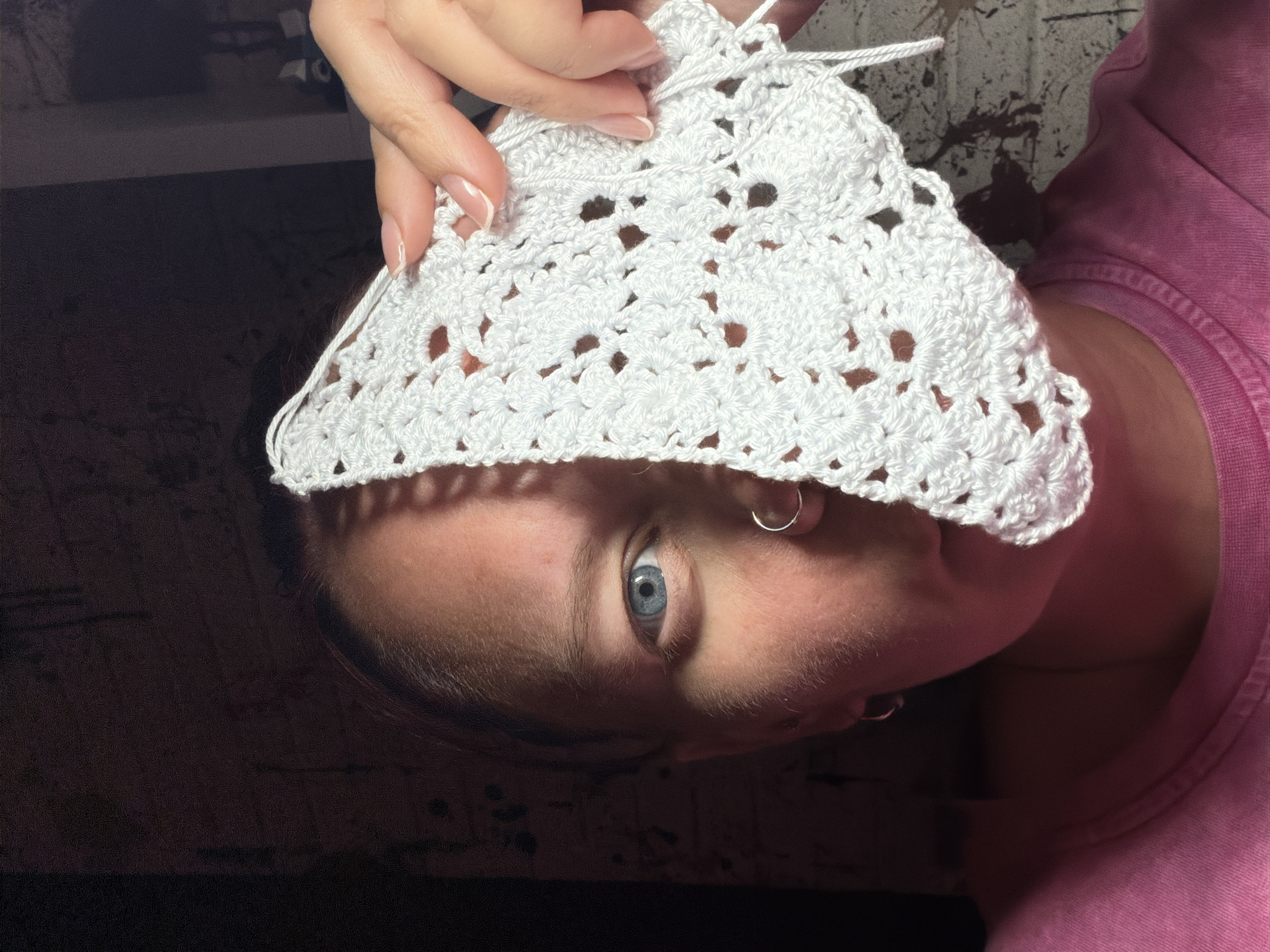 A person with fair skin, light blue eyes, and a silver hoop earring is visible in the frame. The person is wearing a pink shirt and is holding a white crocheted triangular piece of fabric above their face, partially obscuring it. The fabric has a pattern with small holes. The background is a dark surface with a texture pattern, and part of a light-colored brick wall is visible behind the fabric. The image appears to have been taken indoors with mixed lighting conditions. There is no information to support the claim that the location is in the city of Chaikovsky, Russia.