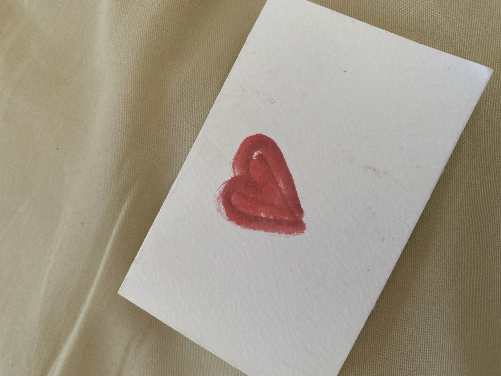 A close-up, overhead shot shows a white, textured rectangular card with a hand-painted red heart on it. The card is placed on a textured, beige fabric surface. The heart appears to be painted with a thick brushstroke, and some smudges of red paint are visible around it. The fabric has a subtle sheen and is likely a tablecloth or upholstery. The image has a shallow depth of field, with the card and heart in sharp focus, and the background slightly blurred. There are no people or other objects visible in the frame. This image is not specific to any location in North Kuta, Indonesia, as it depicts a generic object and setting.