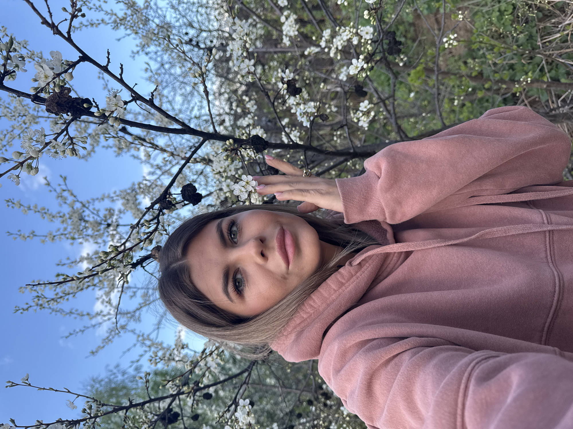 A young woman is posing for a selfie in an outdoor setting. She is wearing a pink hooded sweatshirt and has blonde hair. She is positioned near a tree with white blossoms. The sky is visible in the background, appearing blue with some light cloud cover. The photo is taken from a low angle, looking up at the woman and the tree. The specific location within Chaikovsky, Russia, is not discernible from the image.