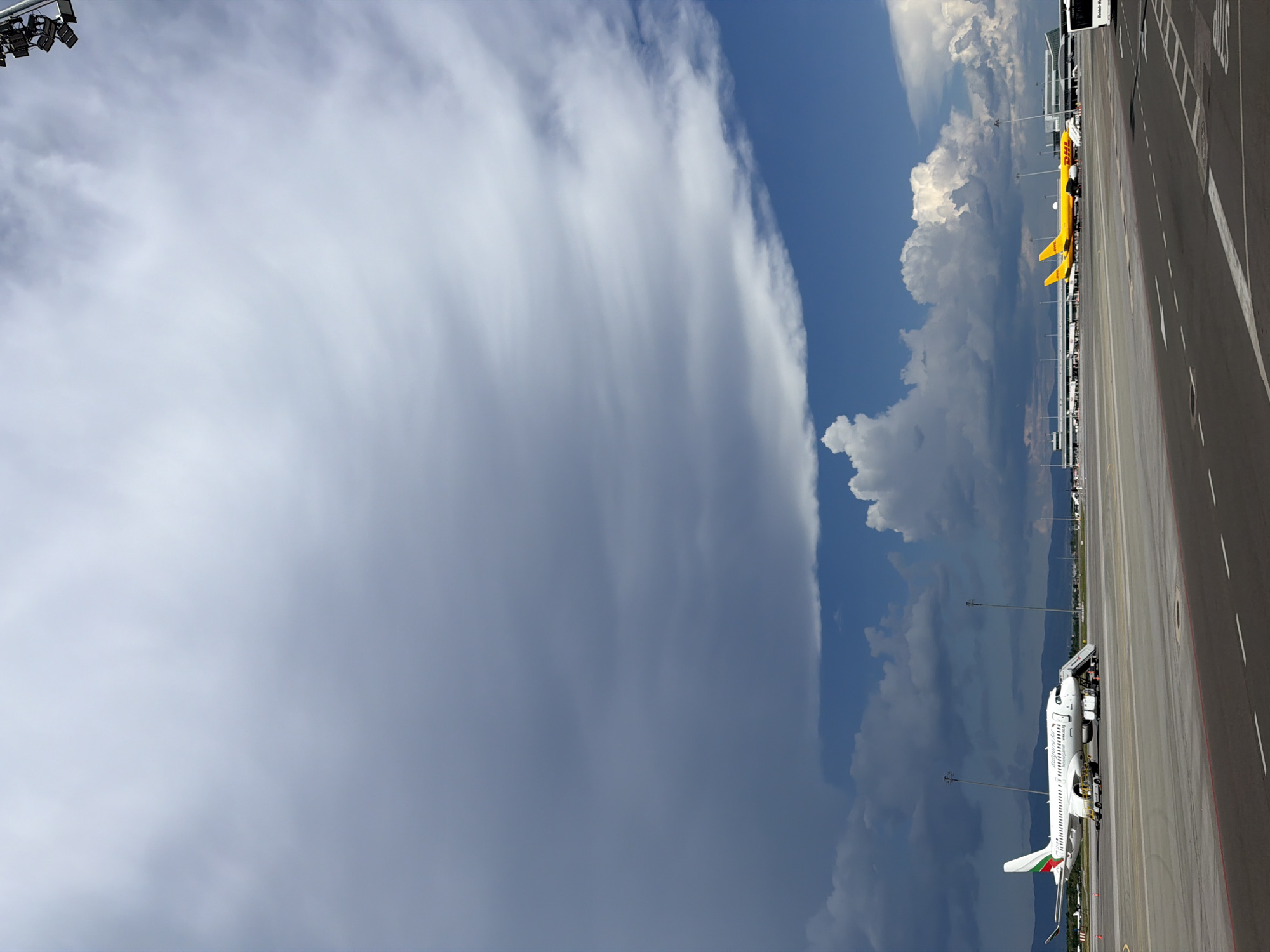 The image depicts a scene at Sofia Airport (SOF), Bulgaria. In the foreground, a tarmac area is visible with runway markings. To the left, a white Bulgaria Air airplane is parked, with a boarding stair extended to its door. Several airport service vehicles are positioned around the aircraft. Further in the background, on the tarmac, a yellow DHL cargo airplane is visible. Beyond the airplanes, the Sofia Airport terminal buildings and other airport infrastructure are present. The sky above is dramatic, with a large expanse of dark, heavy clouds dominating the upper portion of the image, contrasted with patches of bright blue sky and scattered white cumulus clouds. In the distance, a range of mountains is visible under the cloudy sky. The overall scene suggests an airport environment with a significant cloud formation overhead, possibly indicating impending weather changes. There are no people clearly visible in the image.