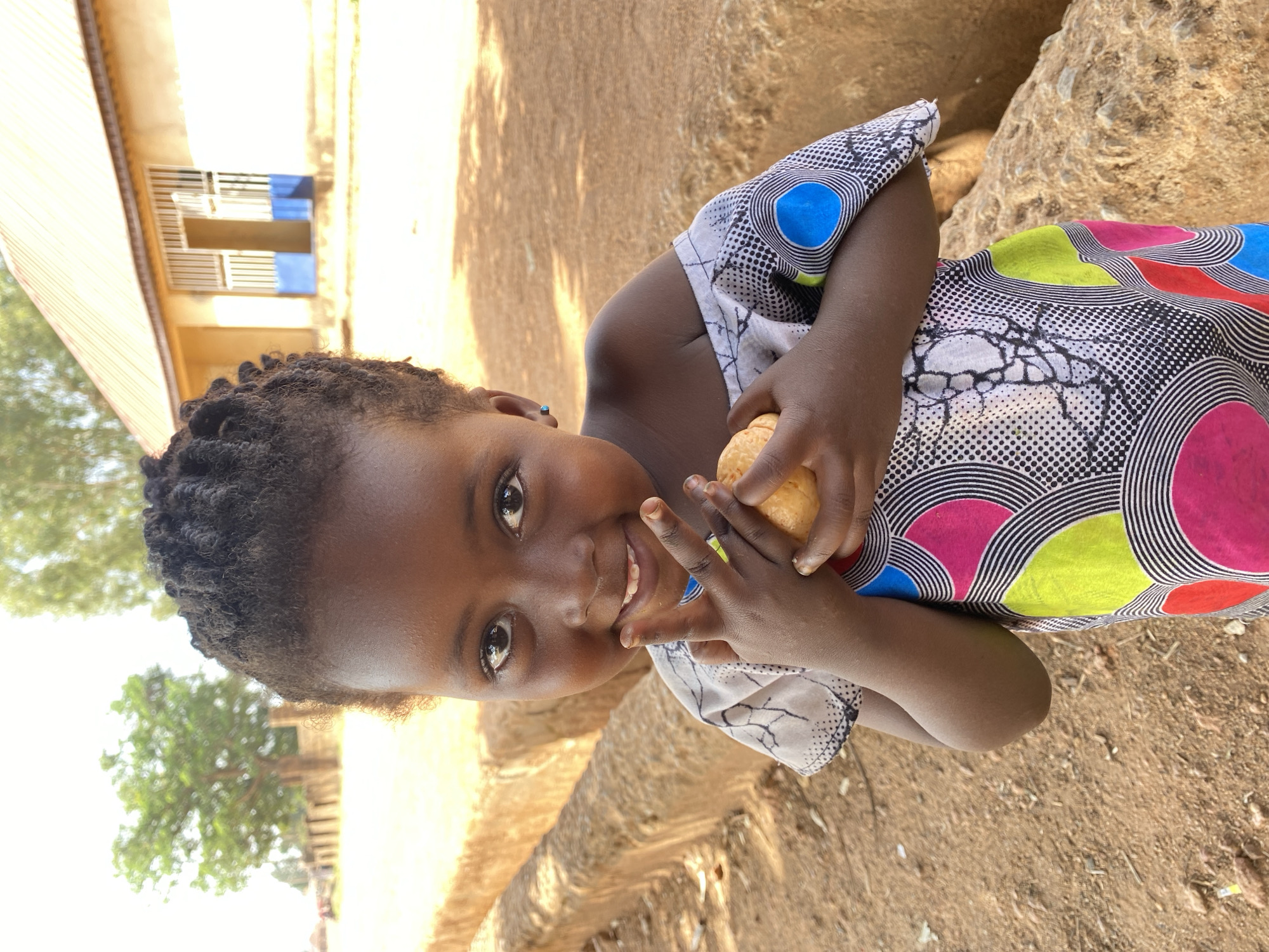 A young, dark-skinned child with braided hair is captured in a vibrant, close-up portrait, looking directly at the camera with a wide, joyful smile. The child is wearing a colorful dress featuring large, stylized circles in shades of pink, green, blue, and red, outlined with black and white stripes, interspersed with patterns of black and white dots. The child holds a light-colored, spherical object, possibly a piece of bread or fruit, in both hands, positioned near their chest.
The scene is outdoors, bathed in bright daylight, indicated by strong sunlight and clear shadows. The background is a mix of light brown, sandy or dirt ground. To the left, part of a light-colored building with barred windows, featuring blue accents, is visible. Further right, there are natural rock formations or uneven terrain. A blurred green tree can be seen in the distant left background. The overall impression is one of happiness and warmth, set in a sunny, possibly rural or semi-urban environment consistent with Kuru Dungus, Nigeria. There is no visible text in the image.