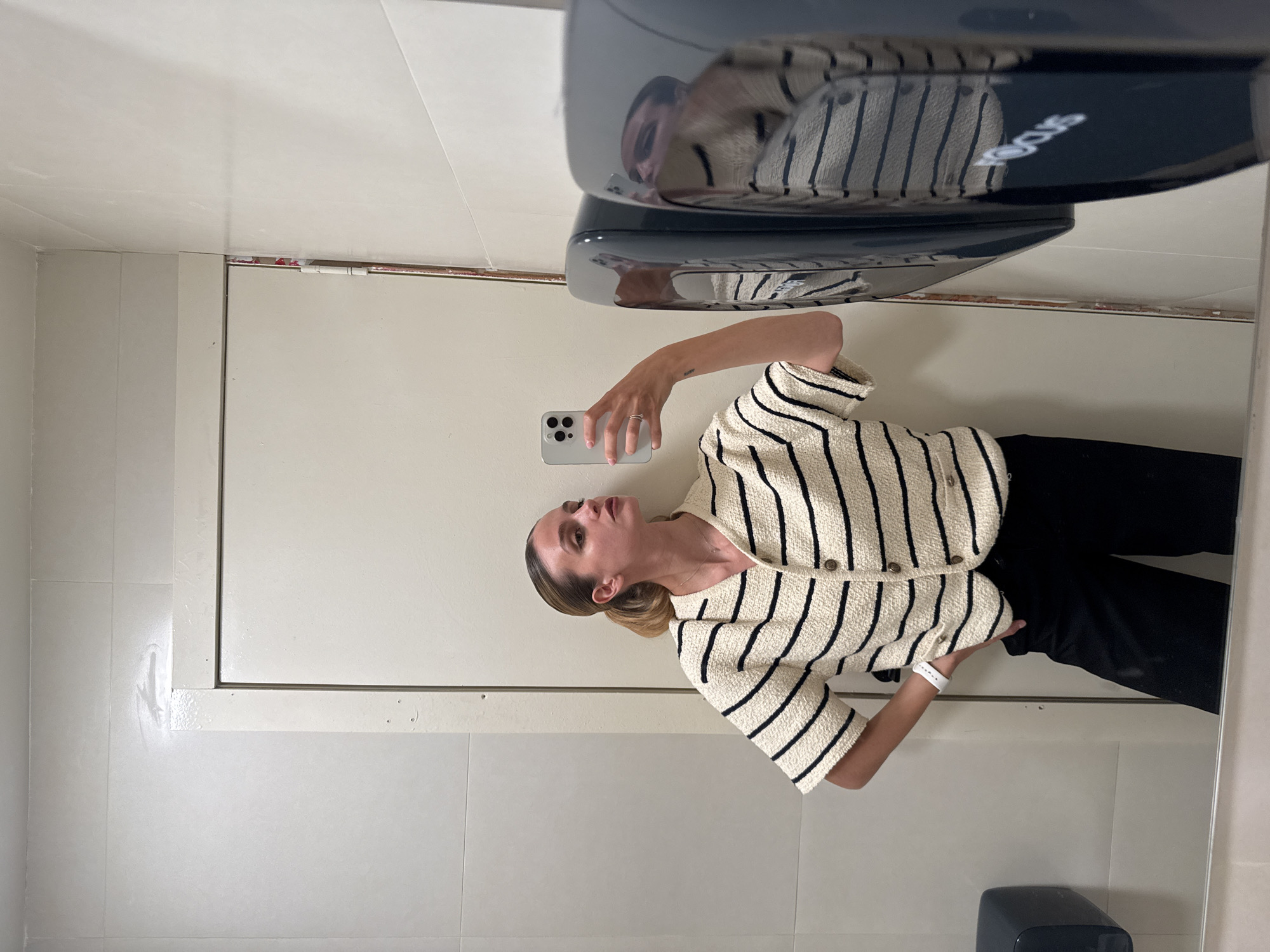 The image captures a woman taking a mirror selfie in what appears to be an indoor public space, possibly a restroom or changing room. The woman, with light skin and light brown hair styled in a bun, is dressed in a cream-colored, short-sleeved cardigan with black horizontal stripes, paired with dark pants. She holds a light-colored smartphone in her right hand, angled to take her reflection. A white wristband is visible on her left wrist, and a silver ring is on her right ring finger. Her gaze is directed towards the phone, with a neutral expression.
The setting features light-colored walls, with the lower sections appearing to be tiled. Above the woman's reflection, a dark gray hand dryer is mounted on the wall. The reflection on the hand dryer reveals part of the word "ECOS" in white lettering, though it is mirrored and partially obscured. To the left of the woman's reflection, a portion of a light-colored door or partition with a metal frame and hinges is visible. At the bottom of the reflective surface, a dark dispenser, possibly for soap, is present. The lighting is even, suggesting an interior environment.