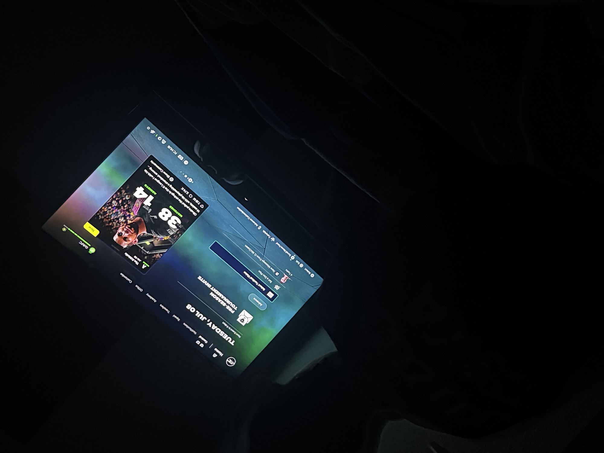 A tilted, dim image shows a screen with a game open, set on Tuesday, July 08. The interface includes options for Squad, Transfers, Academy, and Office. The game's name is difficult to discern but may involve "GrihFC." The screen displays a player graphic with stats: 38 Matches, 14 Assists. Text mentions "Oliveira" and "K League 1." The environment surrounding the screen is dark, with a hint of a textured or fabric surface. There's no evidence of location in Welshpool, United Kingdom.