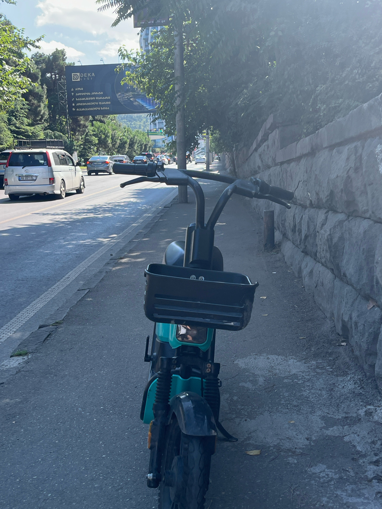 The image depicts a street scene in Tbilisi, Georgia, focusing on a turquoise electric scooter parked on the sidewalk. In the background, a busy road is visible with several cars driving by, including a silver minivan with the license plate "NV-001-RL" and a gray sedan. A large billboard for "DEKA LISI" is prominently displayed on the left side of the road, partially obscured by trees. The right side of the image features a stone wall, suggesting an urban setting. The sky is partly cloudy with bright sunlight, casting shadows on the pavement. The scooter is facing away from the viewer, with its handlebars, basket, and front wheel clearly visible.