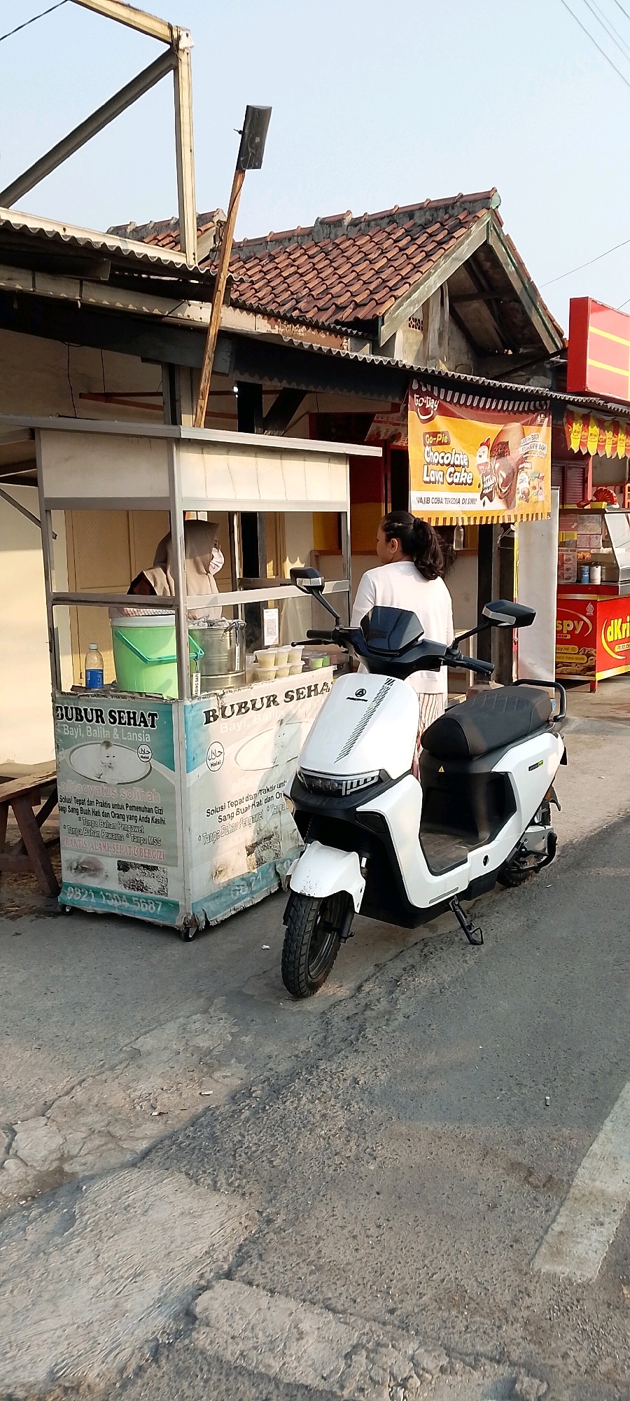 A white electric scooter is parked on a cracked asphalt road in front of a food stall in Satriamekar, Indonesia. The stall, decorated with images of food and text in Indonesian, appears to be selling "Bubur Sehat" (healthy porridge). A woman wearing a headscarf and a face mask is visible behind the counter, serving a customer who is partially obscured by the scooter. In the background, there is a building with a tiled roof and a sign advertising "Go-Pie Chocolate Lava Cake." The scene suggests a typical street-side food vendor operation in Indonesia.