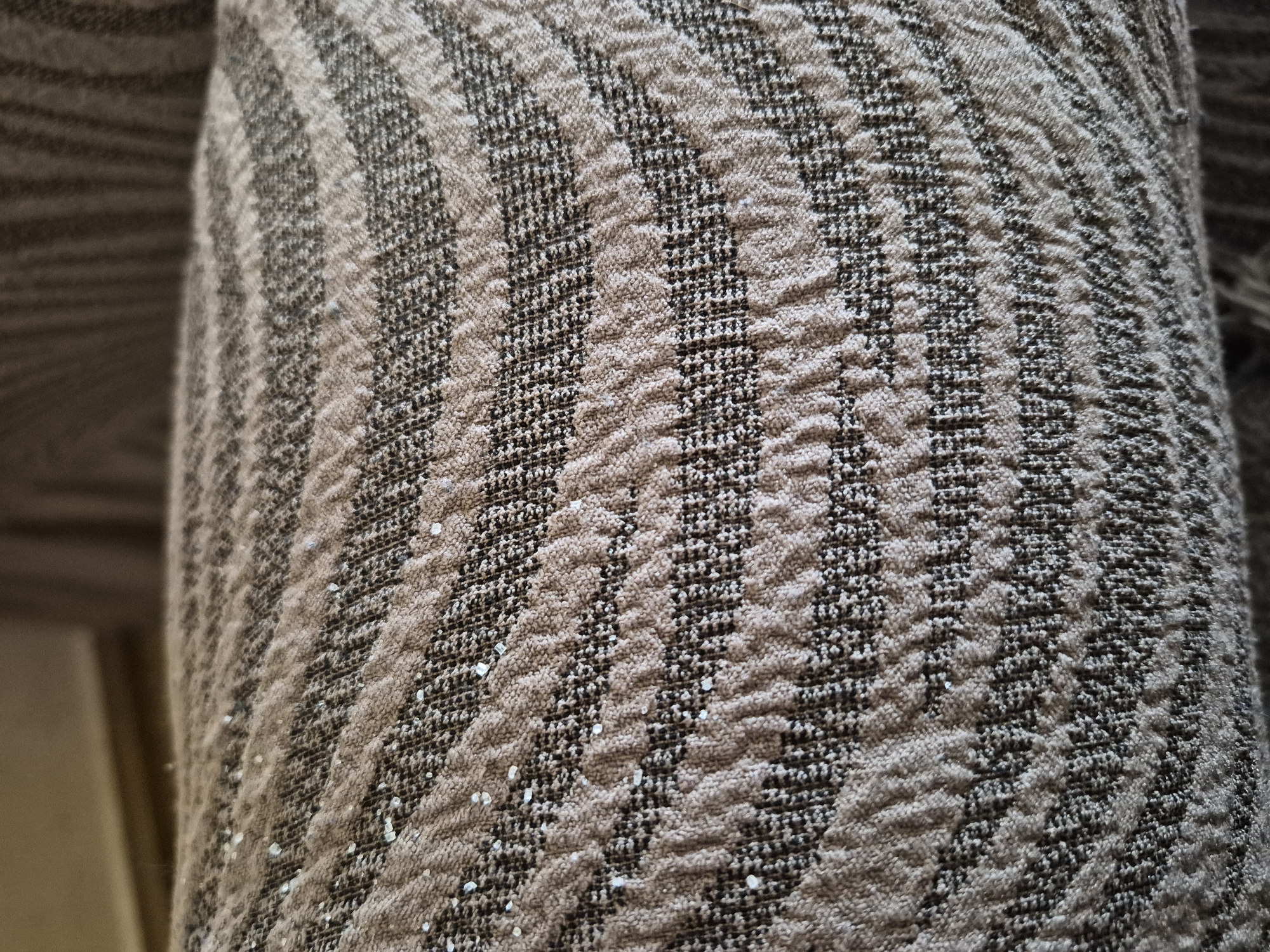 The image is a close-up photograph of a textured fabric, likely upholstery, a rug, or a blanket. The fabric features a prominent pattern of alternating vertical stripes. One set of stripes is a light beige or sandy brown color, characterized by a raised, wavy, or ripple-like texture, giving it a tactile, three-dimensional quality. The other set of stripes is a darker brown or charcoal grey, appearing flatter and more tightly woven with a finer, granular texture.
Scattered across the surface of the fabric, particularly on the lighter textured stripes, are numerous small, irregularly shaped, bright, and somewhat translucent crystalline or glitter-like particles. These particles reflect light, creating subtle glints against the fabric.
The photograph employs a shallow depth of field, with the fabric in sharp focus in the foreground, while the background is softly blurred and indistinct, suggesting an indoor setting. No people or specific activities are depicted. The lighting appears to be ambient indoor light. There is no visible text in the image. The general appearance suggests a worn or well-used textile.