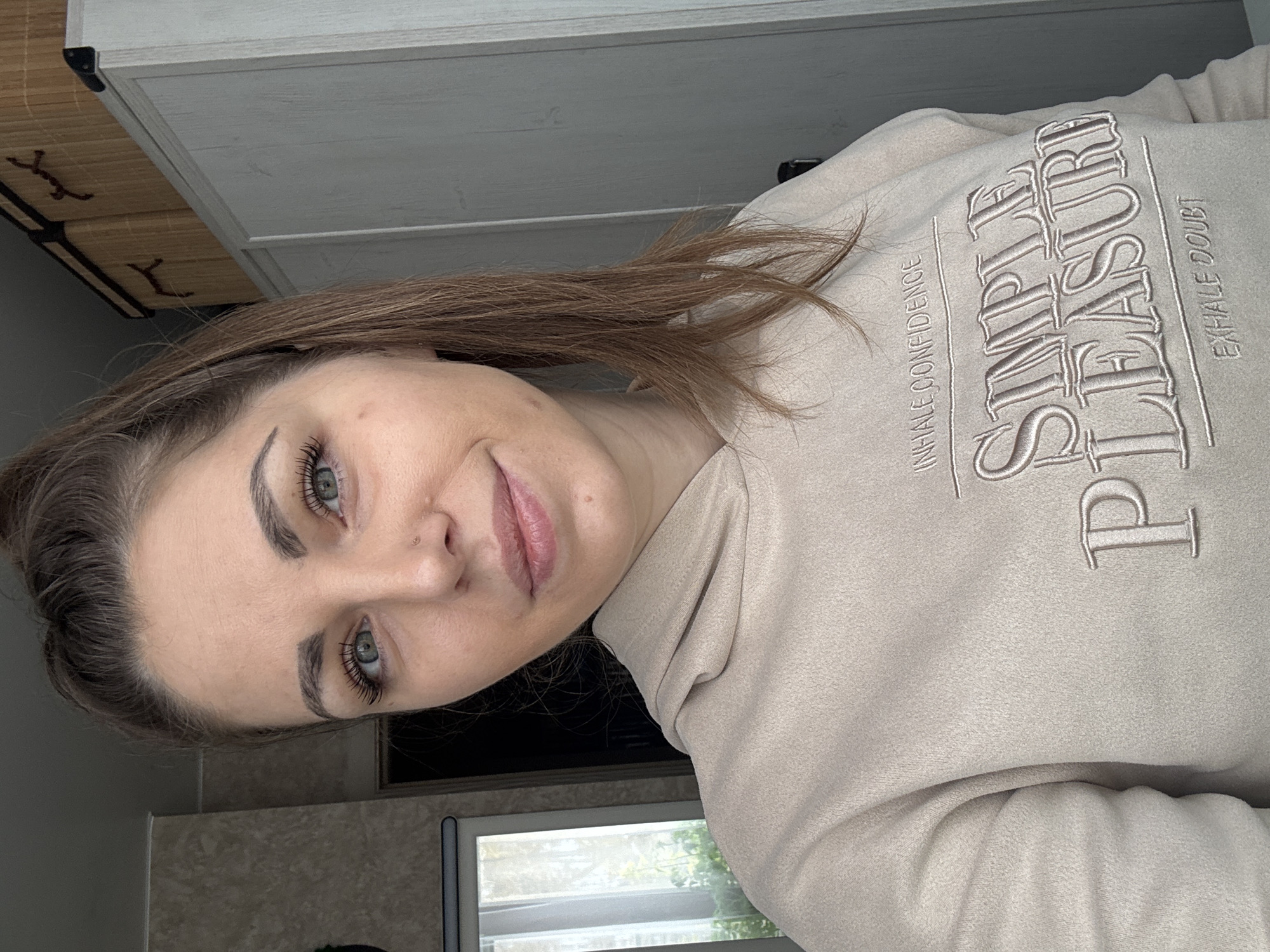 The image, rotated 90 degrees counter-clockwise, features a young woman in an indoor setting, likely in Раздолье, Russia. She is positioned in the center-right of the frame, looking towards the viewer with a slight, content smile and a gentle head tilt. Her light brown hair is visible, falling over her shoulders. She has light-colored eyes, defined eyebrows, and prominent eyelashes enhanced with mascara. A small nose stud adorns her left nostril, and a mole is visible on her right cheek.
She is wearing a light beige or cream-colored hooded sweatshirt with embroidered text on the front. The most prominent phrase reads "SIMPLE PLEASURE" in large, stylized letters. Below "SIMPLE," smaller text states "INHALE CONFIDENCE," and below "PLEASURE," it reads "EXHALE DOUBT."
The background suggests a room during the daytime, indicated by natural light streaming through a window visible on the lower right, revealing green foliage outside. To the upper left, a light-colored wooden cabinet or shelf is present, topped with two distinct woven storage boxes with dark bindings and stylized embellishments. A dark countertop or surface runs below the window. The overall impression is one of calm confidence.