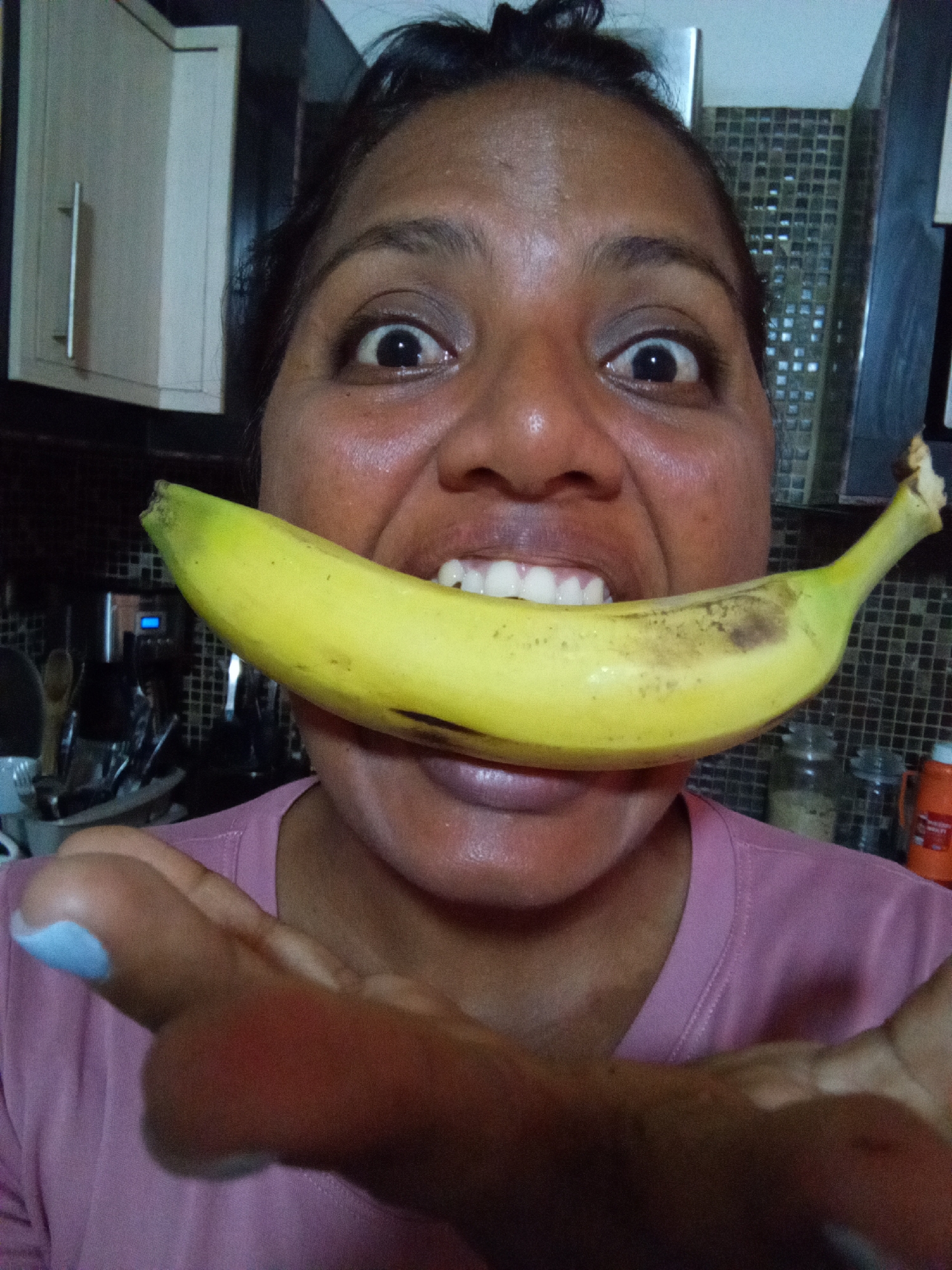 A woman in a pink t-shirt holds a banana in her mouth and extends her hands towards the camera. She has wide eyes and is smiling with her teeth showing. She appears to be in a kitchen, with cabinets and a tiled backsplash visible in the background. Kitchen utensils and jars are visible on the counter. The location is Unknown, Unknown.