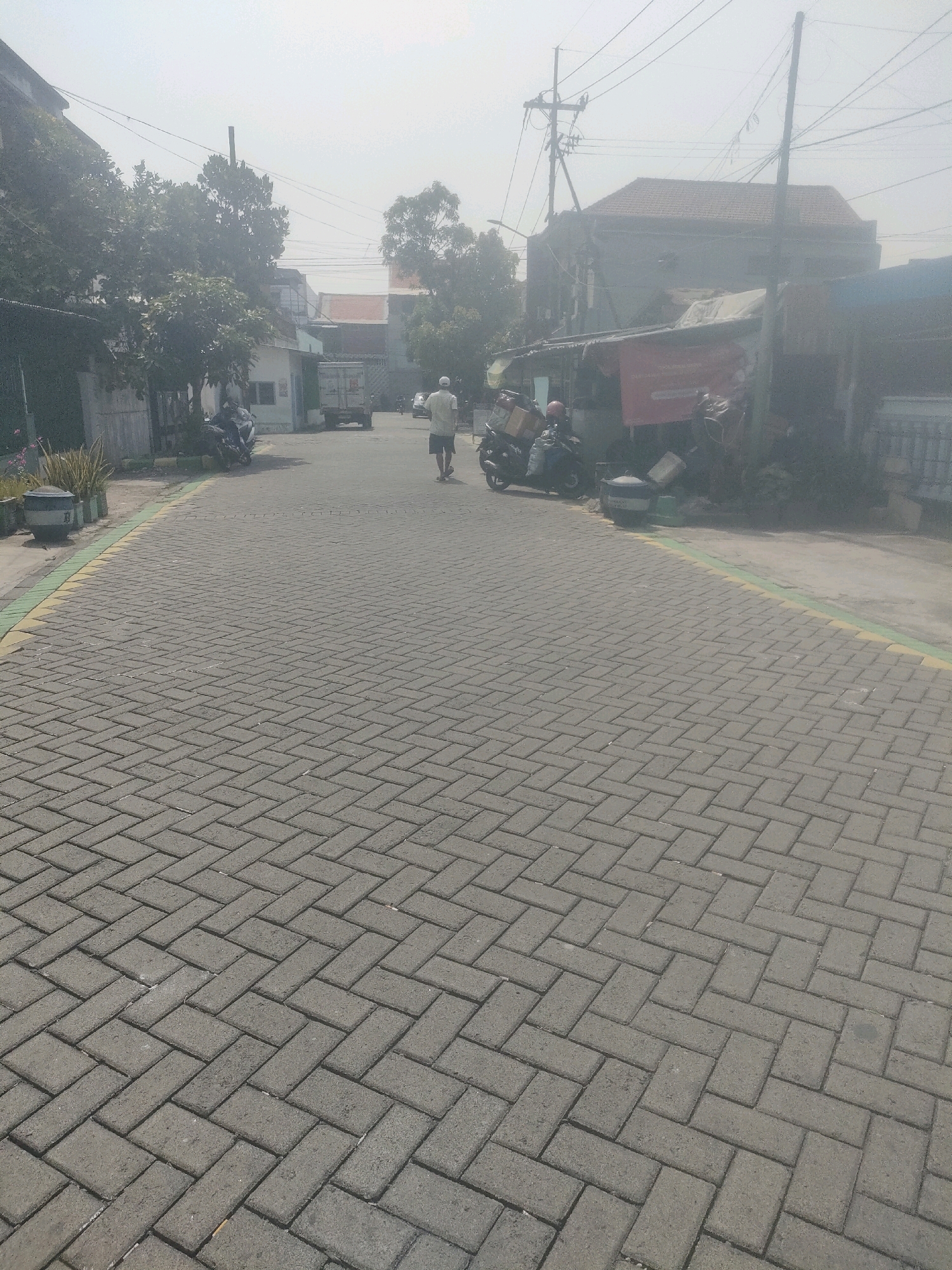 The image depicts a street in Surabaya, Indonesia, paved with interlocking concrete blocks laid in a herringbone pattern. The street is bordered by a yellow and green line on the left side. On the right side of the street, there are buildings, some with red tiled roofs, and a red banner. Several parked motorcycles are visible, one with delivery boxes. A man wearing a light-colored shirt and shorts is walking away from the camera down the street. In the background, a white truck is parked. The scene is hazy, suggesting it may be a hot or dusty day. Overhead, electrical wires are strung across poles. Trees are present along the sides of the street and behind some of the buildings.