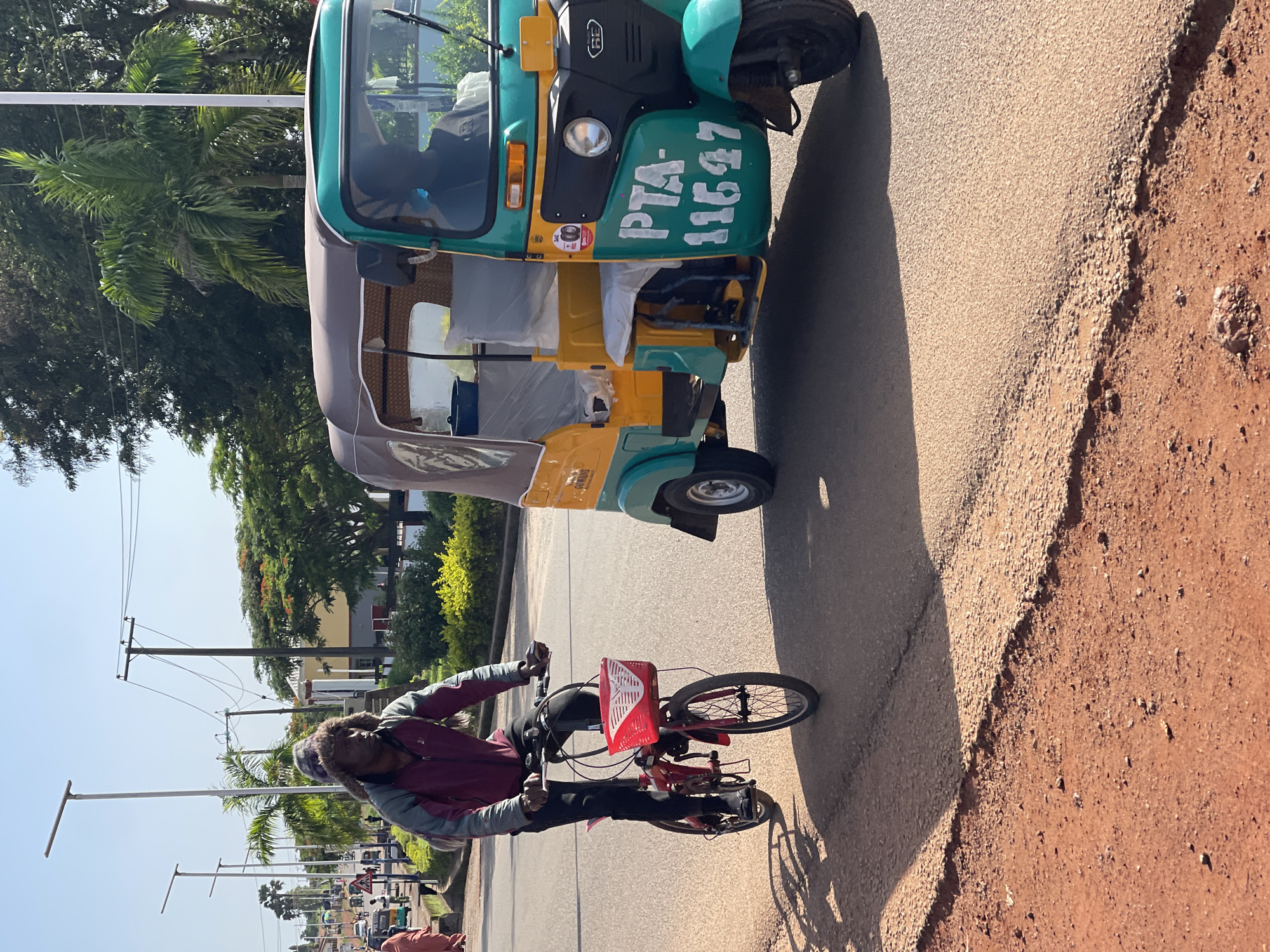 A man is riding a red folding bicycle on a paved road in Jos, Nigeria. He is wearing a dark jacket and a furry hat. Next to him, a yellow and turquoise auto-rickshaw is driving on the same road. The auto-rickshaw has the license plate "PTA-11647" painted on its side. The road has a red dirt shoulder. In the background, there are palm trees, other trees, buildings, and streetlights. The sky is clear and blue.