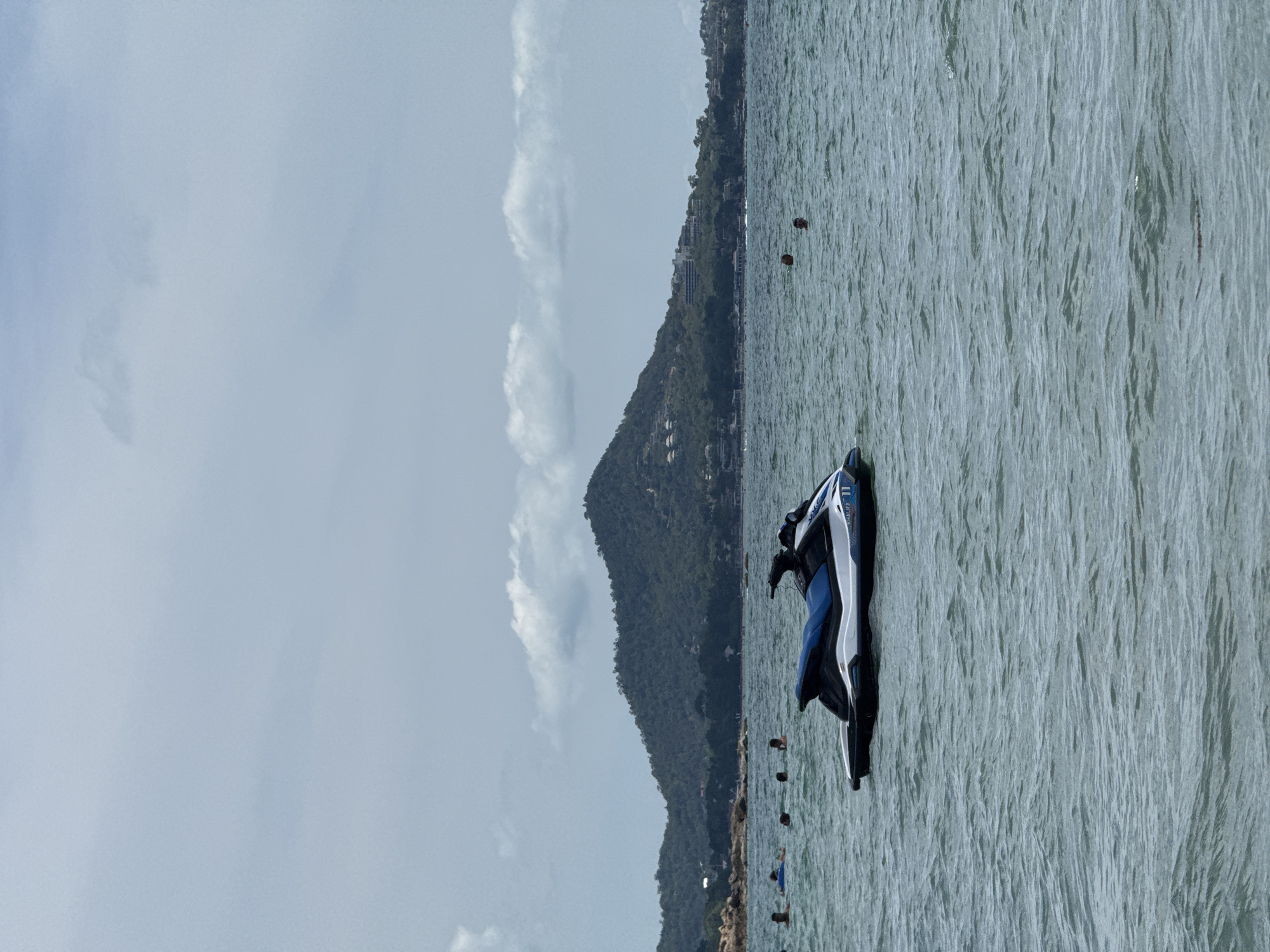 In the city of Ko Samui District, Thailand, a jet ski, predominantly white with blue accents, floats on the surface of a choppy, grey-blue sea. The jet ski displays the number "11" and the word "KHALIFA" on its side. In the background, a large, verdant hill covered in trees and dotted with buildings rises from the coastline. Several people are visible in the water, some swimming and others with their heads above the surface, dispersed across the scene. The sky is overcast with scattered clouds.