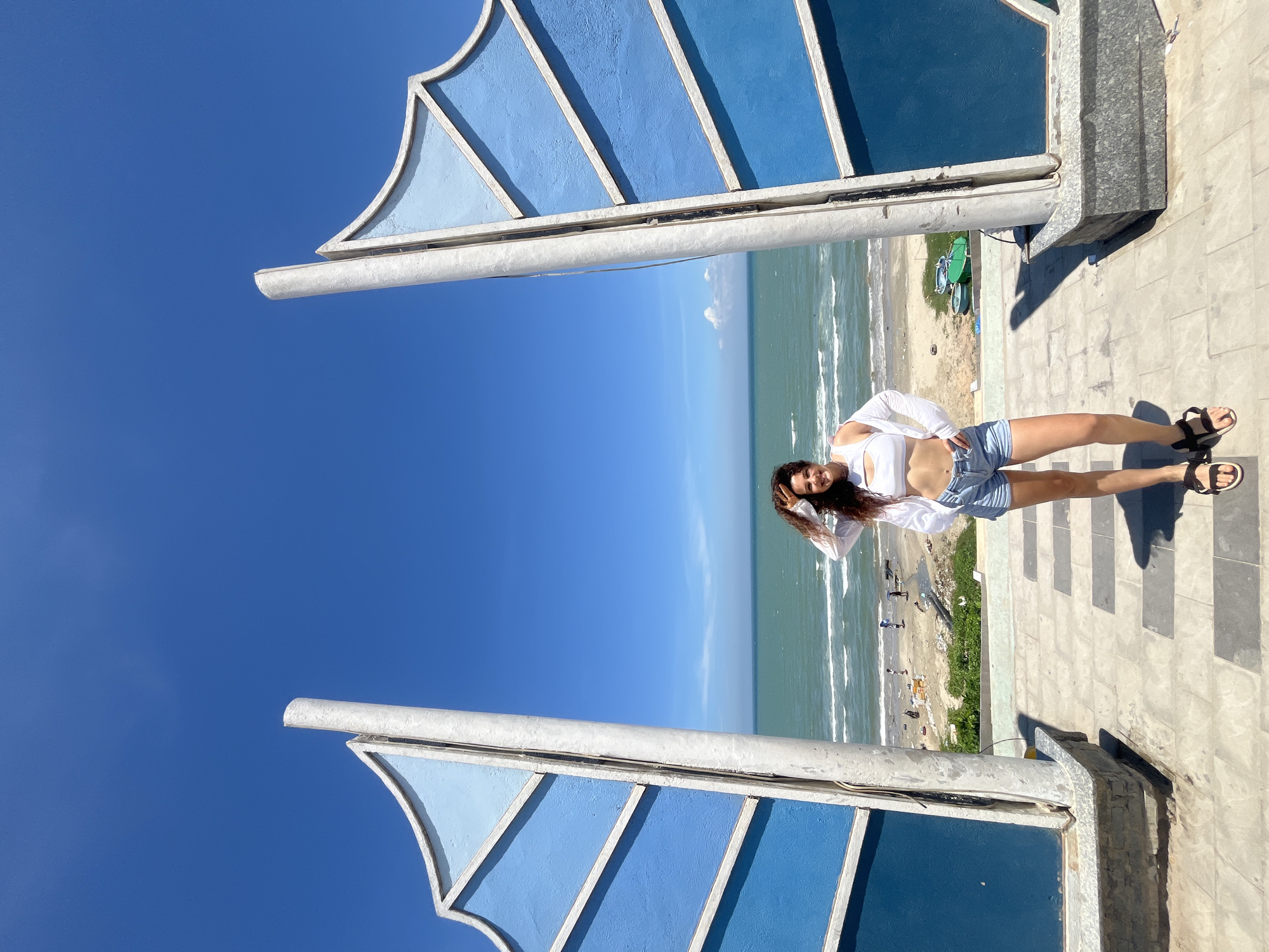 A young woman stands on a paved walkway with a view of the ocean. She is wearing a white crop top, a white open shirt, and denim shorts. Her hand is raised to her head, and she is smiling at the camera. Behind her, the ocean stretches to the horizon under a clear blue sky. Two large, blue, wing-shaped structures frame the scene on either side. The walkway leads down to a sandy beach where a few people are visible, along with some boats. The location is Mũi Né, Vietnam.