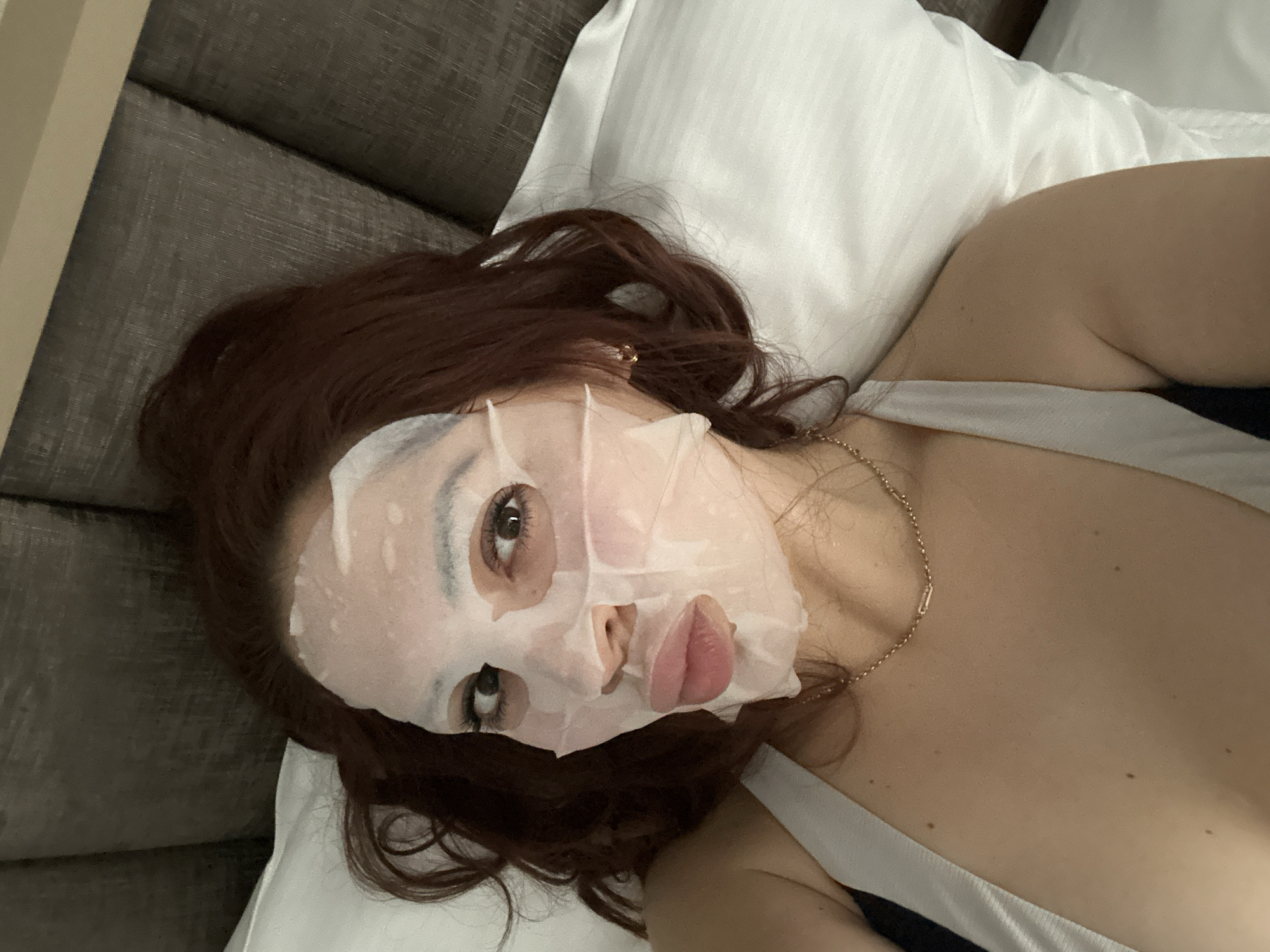 A woman with auburn hair, wearing a white sheet mask on her face, is lying on a white pillow in a bed with a gray headboard. She is wearing a gold necklace and a white tank top. Her location is not discernible from the image alone.