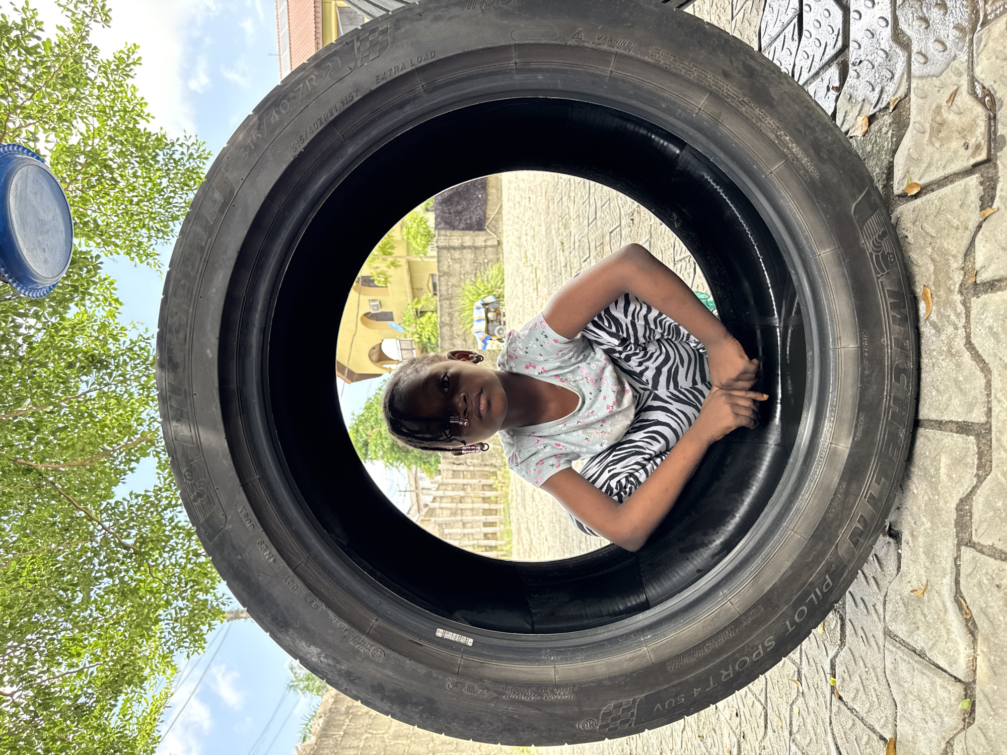 A young dark-skinned child, likely a girl, is playfully captured looking directly at the viewer from within a large black car tire. She is wearing a light-colored short-sleeved top with a subtle floral pattern and distinctive black and white zebra-print pants, and has braided hair with small earrings. Her expression is calm as she crouches or sits inside the tire.
The tire is a "MICHELIN PILOT SPORT 4 SUV," with visible text detailing its size as "315/40ZR21 115Y" and further markings such as "EXTRA LOAD," "DOT AMES 008X," and a "WARNING."
The setting is outdoors on a bright, sunny day, evidenced by the clear blue sky with scattered white clouds and strong illumination. Through the tire's opening, light-colored buildings, possibly with stairs, and abundant green tree foliage are visible in the background. Around the tire, textured paved ground, consistent with interlocking pavers, and more green leaves are present, along with a partial view of a blue plastic object, possibly a bucket or lid. The scene suggests a residential or semi-urban environment in Ado, Nigeria. The image is presented with a 90-degree clockwise rotation from a standard upright viewing orientation.