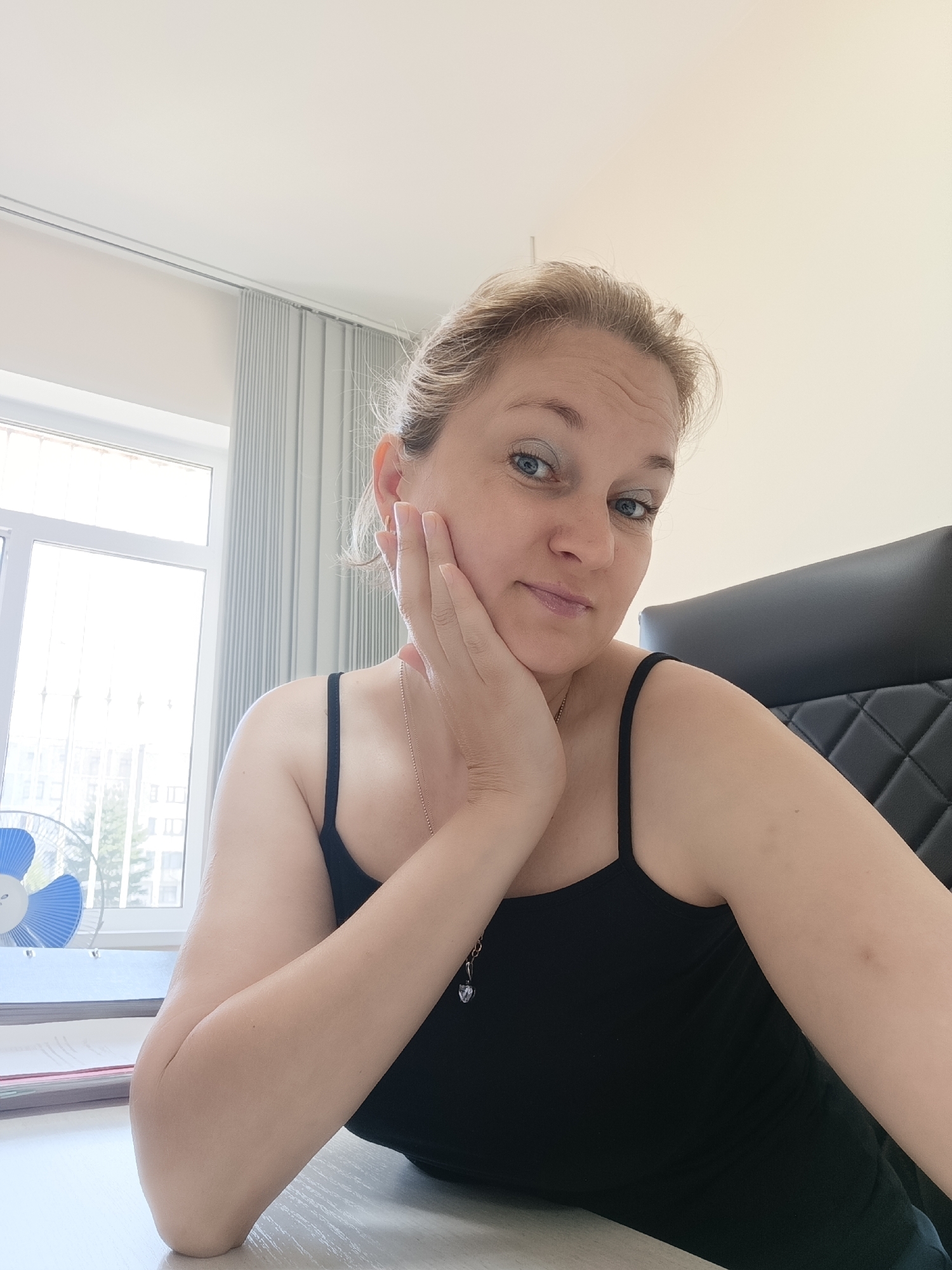 A fair-skinned woman with blonde hair pulled back is seated at a desk in what appears to be an office. She is wearing a black tank top and has her right hand resting on her cheek. Her left arm is bent at the elbow and resting on the desk. She is looking directly at the camera with a neutral expression. The background includes a window with vertical blinds, a blue and white fan, and a black office chair. The scene is indoors and appears to be during daylight hours. There is no information about the specific location within Марковский, Russia.