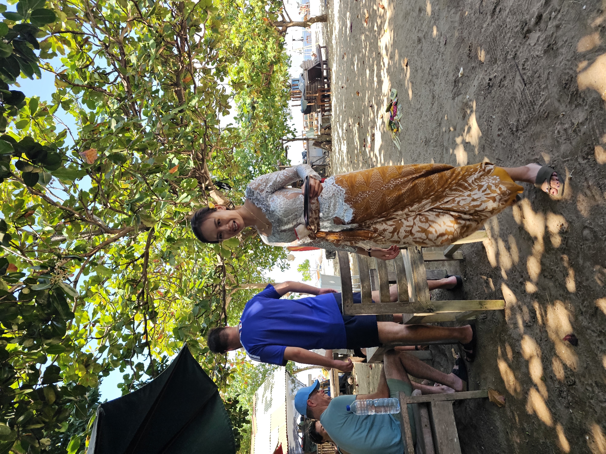 A woman in traditional Balinese attire stands on a sandy beach in Denpasar, Indonesia, holding a tray of food. She is wearing a white lace top and a golden sarong with an intricate pattern. Her hair is pulled back, and she is smiling at the camera. Behind her, several people are seated at wooden tables under the shade of trees. The trees are lush and green, with dappled sunlight filtering through the leaves onto the sand. In the background, lounge chairs and other beach furniture are visible, suggesting a resort or restaurant setting. The scene evokes a sense of tropical leisure and cultural tradition.