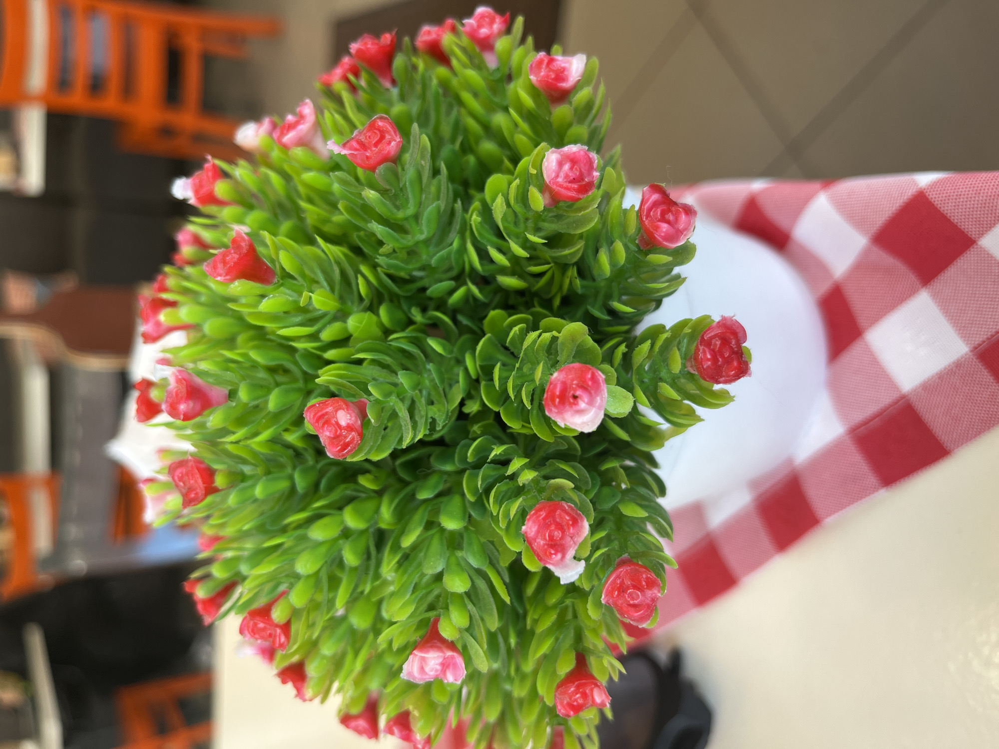 The media file presents a high-angle, slightly rotated shot of a small, artificial potted plant featuring bright green leaves and numerous miniature red and pink rosebuds. The plant occupies the majority of the left and central frame. The leaves are densely packed, appearing to be made of textured plastic, while the small, budding roses have a slightly waxy, artificial finish, predominantly red with some lighter pink accents.
The plant rests in a white pot or container, which is partially visible towards the right. Beneath and to the right of the plant, a red and white checkered tablecloth is prominent, displaying a classic gingham pattern. The background beyond the plant and tablecloth is significantly blurred, indicating a shallow depth of field. Within this blurred background, indistinct orange, ladder-like structures and darker, abstract shapes are visible, suggesting furniture or architectural elements in an indoor setting.
There are no people or specific activities depicted. The scene is static, focusing on the decorative plant. No text is visible in the image. The setting appears to be indoors, potentially a cafe, restaurant, or home environment in Ulyanovsk, Russia. The lighting suggests ambient indoor conditions.