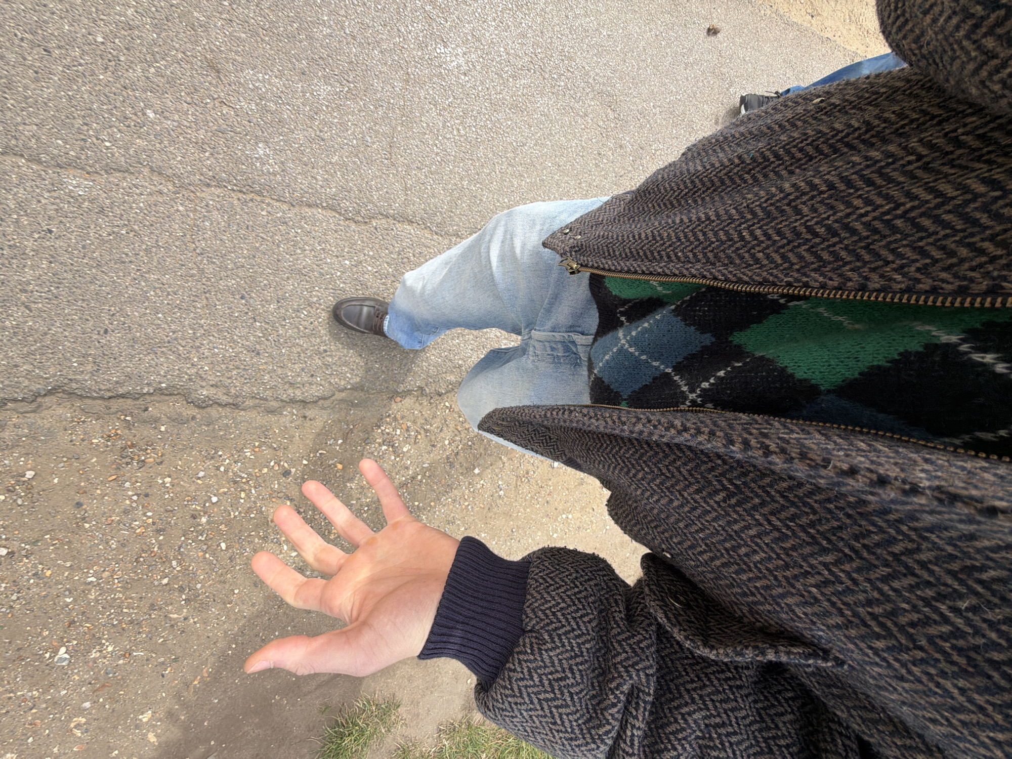 The image is a first-person view of a person standing on a concrete surface, possibly a sidewalk or road, in an outdoor environment. The person is wearing light blue denim jeans, brown leather shoes, and a dark brown herringbone pattern jacket over a green and blue argyle sweater. The person's left hand is outstretched towards the left side of the frame. The surface underfoot is grey concrete with visible cracks and patches of discoloration. There is green grass visible in the bottom right corner of the frame. No specific information suggests the location being London, United Kingdom.