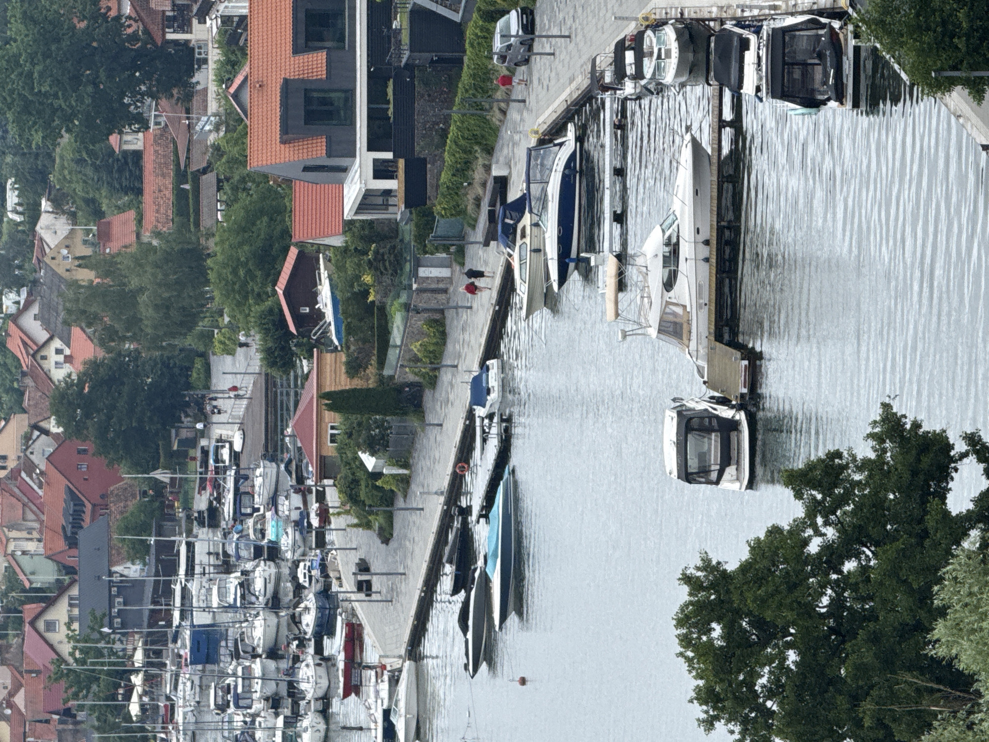 The image depicts a harbor in Mikołajki, Poland, on a cloudy day. The water is calm, with several boats docked along a paved walkway. The boats vary in size and type, including yachts, speedboats, and smaller motorboats. Some boats have canvas covers, while others are open. A few people are visible walking along the walkway, and one person is standing near a silver car.
In the background, a town with red-tiled roofs and multi-story buildings rises above the harbor. Lush green trees are interspersed among the buildings and along the shoreline. The overall scene is one of a well-maintained recreational harbor with a picturesque town backdrop.