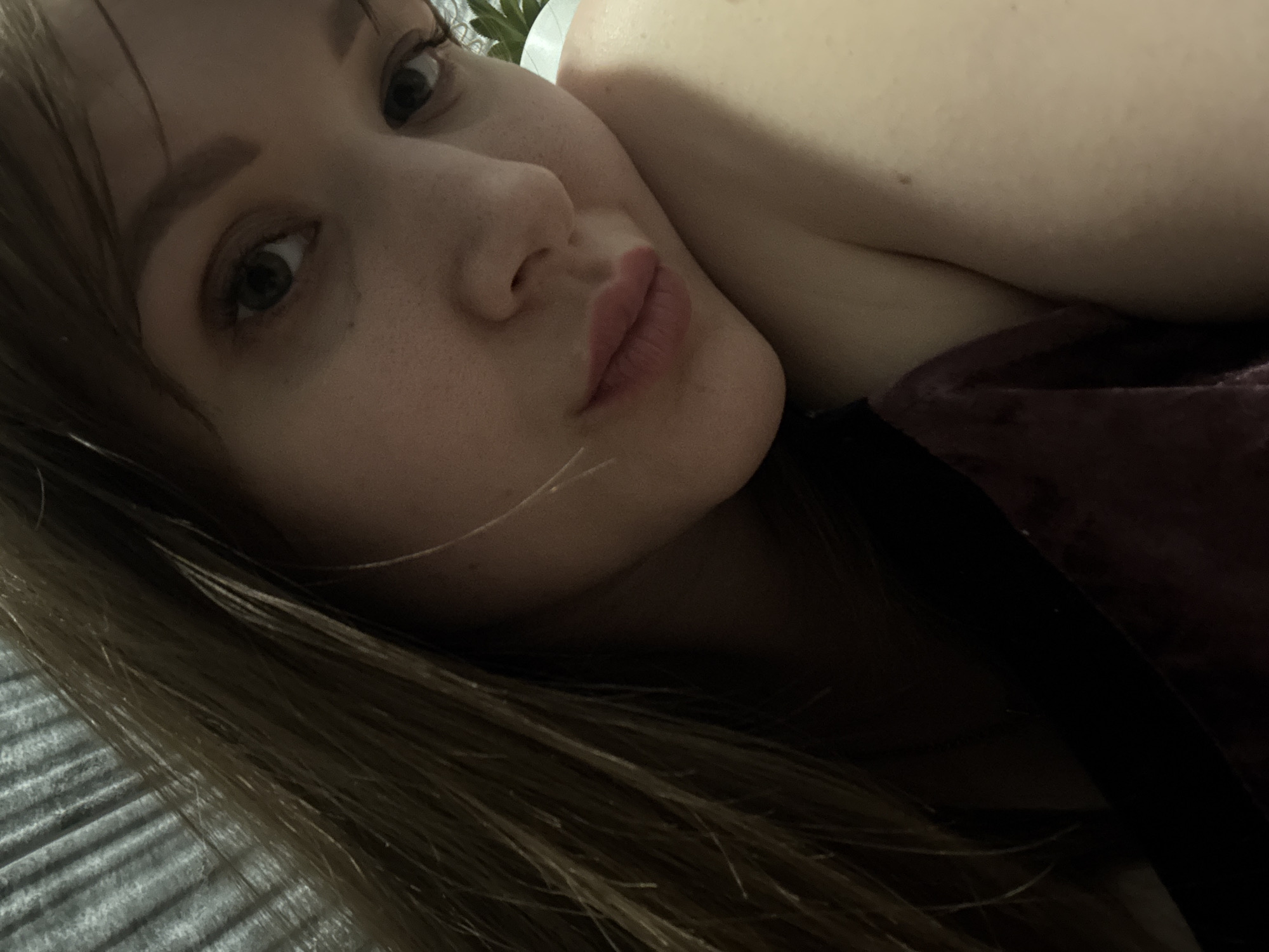 A close-up, angled shot of a woman lying down, likely indoors. She has fair skin, light brown hair with blonde highlights, and natural-looking makeup. The background is out of focus, but suggests a bed or upholstered surface. Her clothing is partially visible, appearing to be a dark-colored, possibly velvet top. The location is not discernible from the visual data.