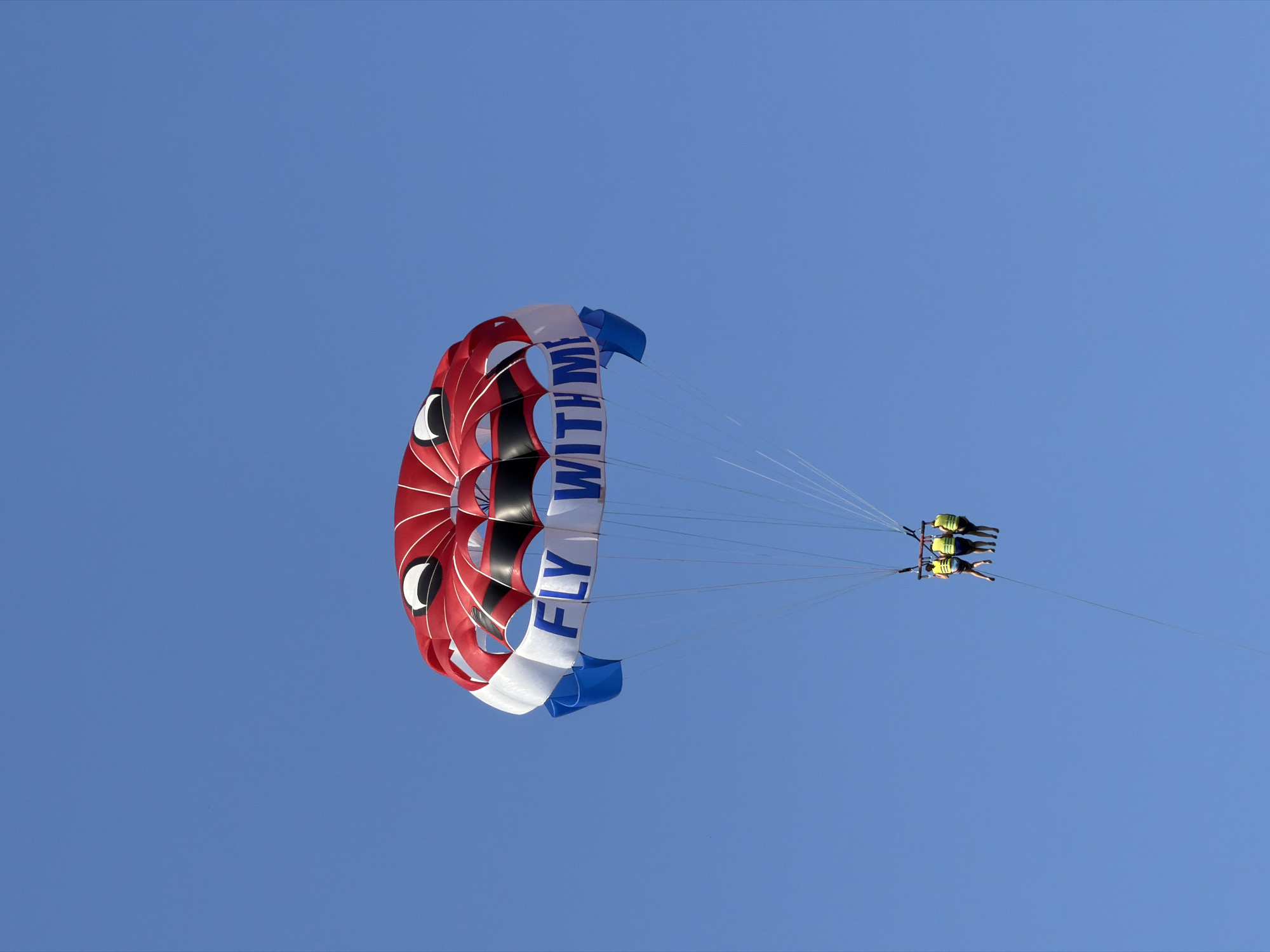 A bright red, white, and blue parachute, with the words "FLY WITH ME" written in blue letters on its white panels, soars through a clear blue sky. Attached to the parachute are three people wearing bright yellow life vests, suspended in the air. The individuals appear to be enjoying a parasailing adventure. The scene is set against a uniform, cloudless sky, suggesting good weather conditions for outdoor activities. The image is cropped to focus on the parachute and the people, with no visible city or ground features.