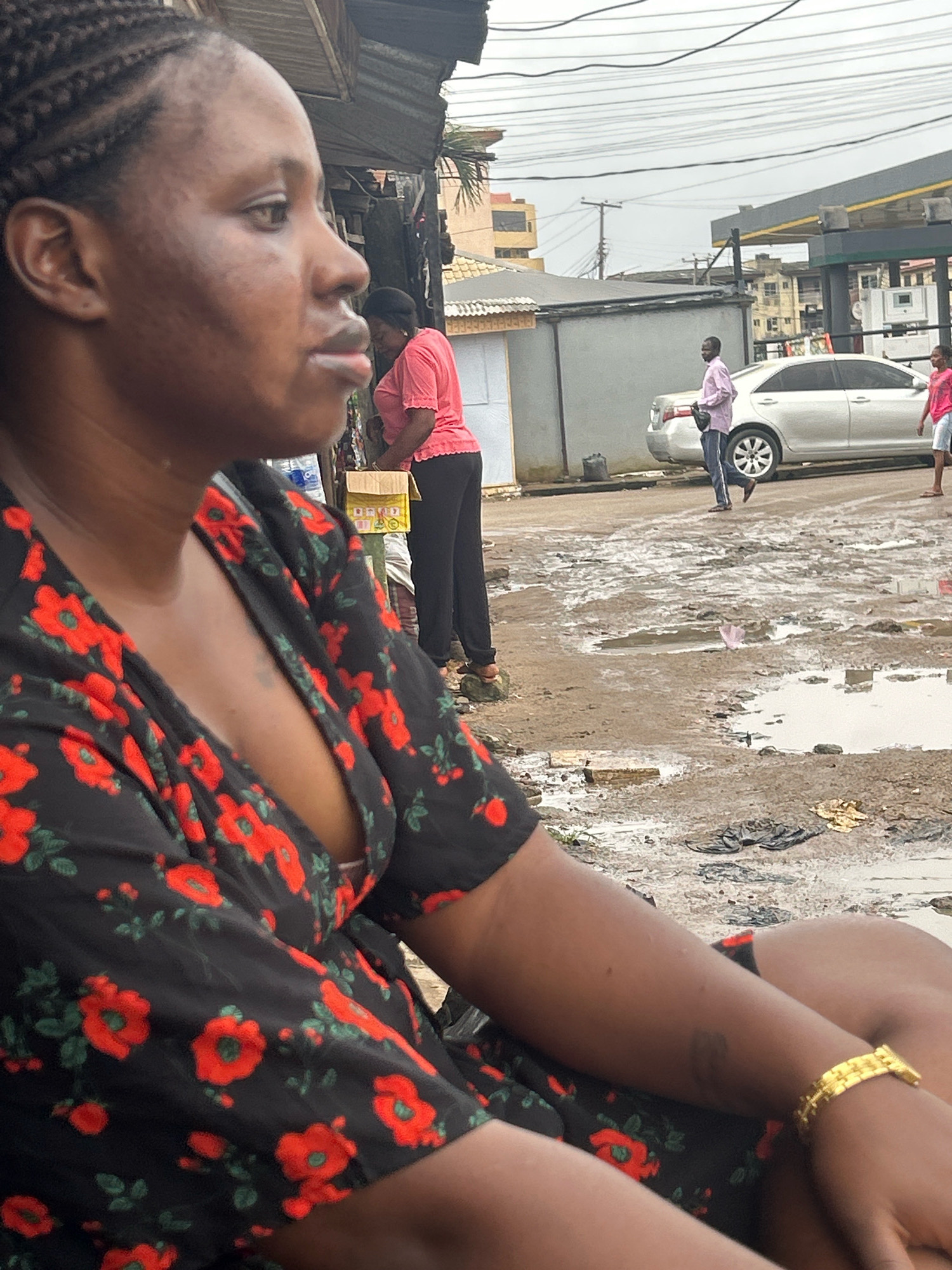 The image depicts a side-profile view of a Black woman with braided hair, seated outdoors in what appears to be a street or market area in Ikotun, Nigeria. She is wearing a black dress with a red floral pattern and a gold bracelet on her right wrist. Her arms are crossed, and she is looking to her left.
In the background, there are other individuals, a silver car, buildings, and what looks like a gas station canopy. The ground is muddy with several puddles, suggesting recent rain. Power lines are visible overhead. The overall environment suggests an urban setting that is experiencing inclement weather.