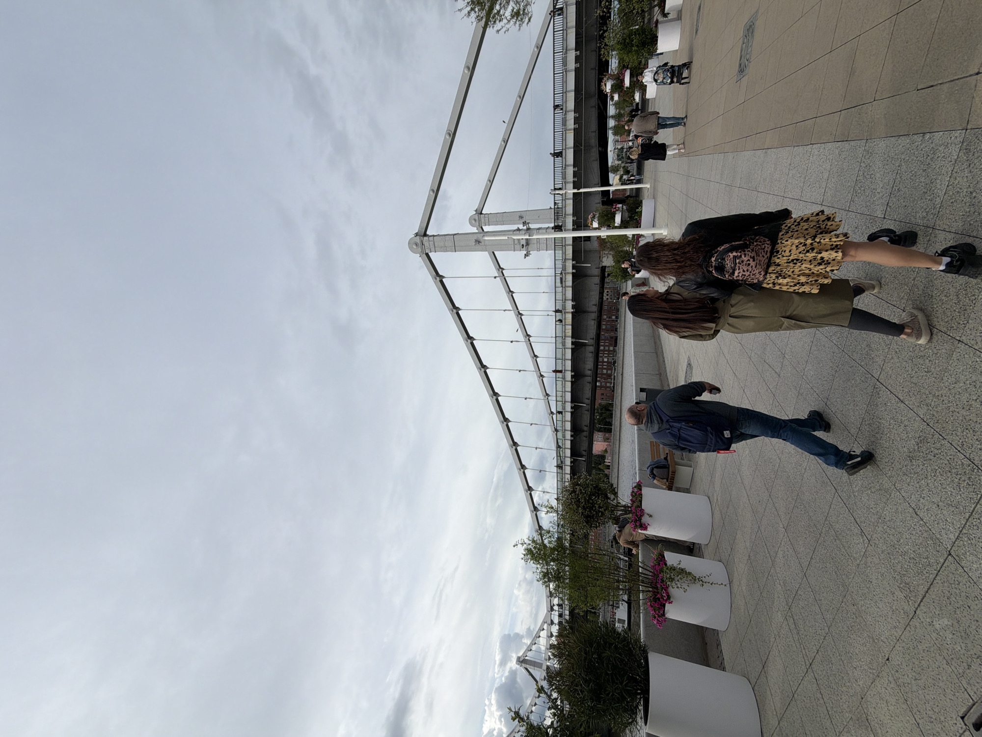 The image captures a street scene in Moscow, Russia, with a view of a bridge featuring a white metal support structure. The sky is cloudy. A man in jeans and a blue jacket walks on the sidewalk. Two women stand nearby, one in a leopard-print skirt and the other in an olive-green coat. There are white planters with green bushes and flowers along the sidewalk. Other people are visible in the background.