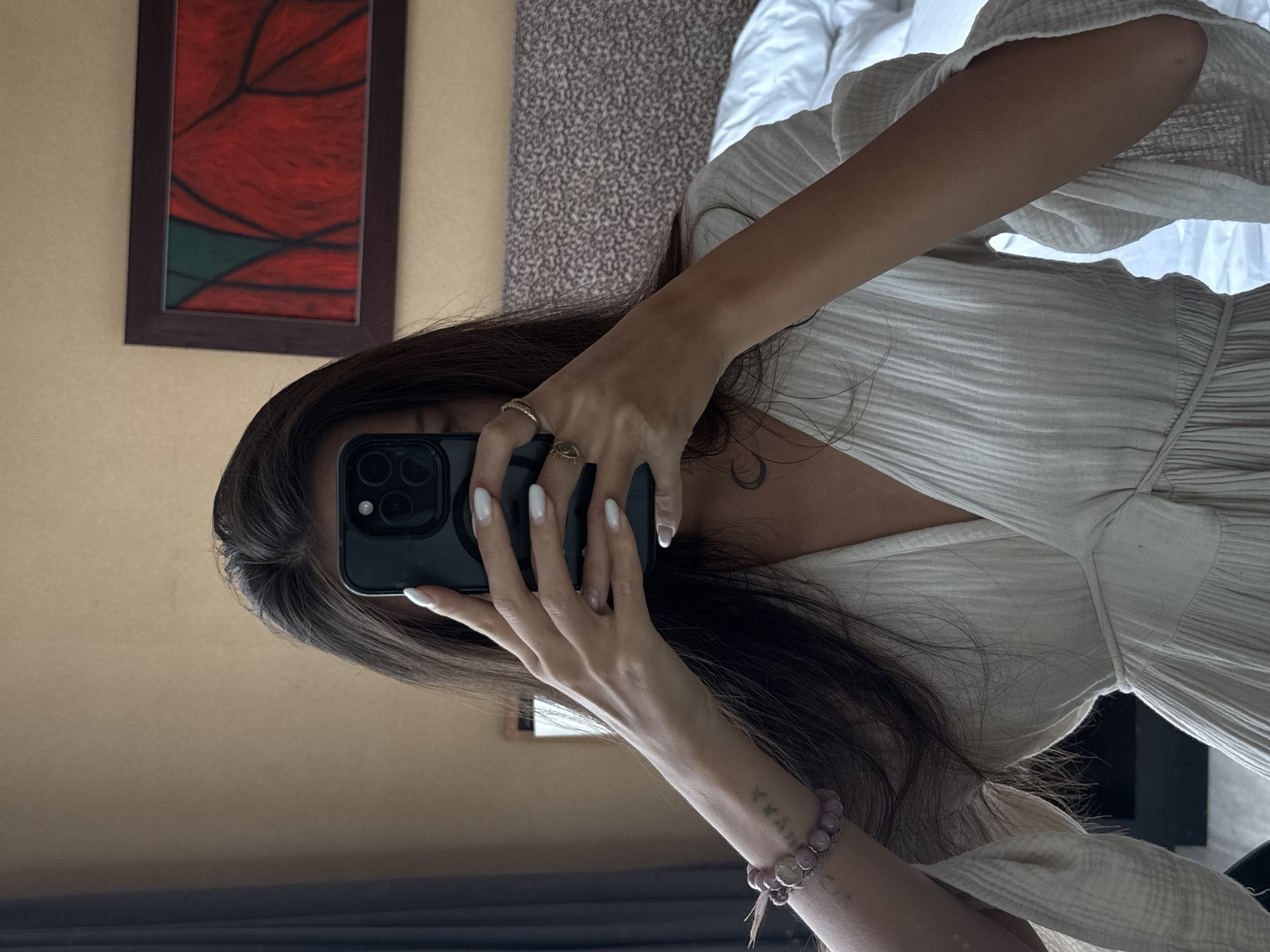 The image shows a person taking a mirror selfie in a hotel room. The person is holding a smartphone in front of their face, obscuring their features. They are wearing a light-colored, textured dress with wide sleeves. On their left wrist, they are wearing a beaded bracelet. There is a subtle tattoo visible on their left forearm. In the background, a framed abstract painting with red and green elements hangs on a tan wall. A headboard of a bed, covered with a patterned fabric, is also visible, along with a corner of a white duvet. The setting appears to be a hotel room, likely in Bangkok, Thailand, given the provided context.