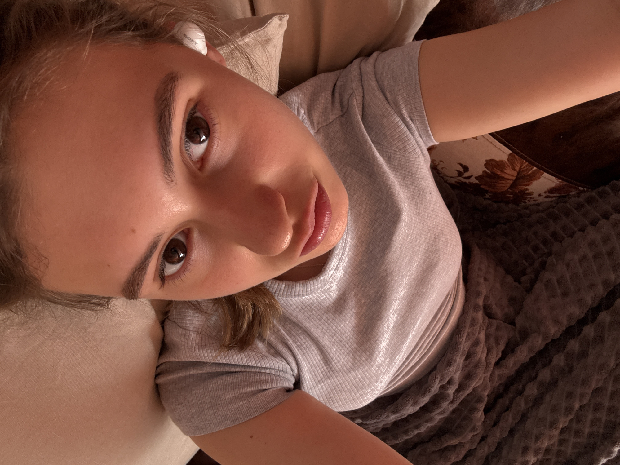 A young woman lies at an angle on a light-colored cushion, partially covered by a brown textured blanket. She wears a gray ribbed t-shirt and has a white Sony wireless earbud in her left ear. Her brown eyes are visible, and her lips appear glossy. The background includes what seems to be a floral-patterned surface. The location in Panagyurishte, Bulgaria, is not discernible from the image.
