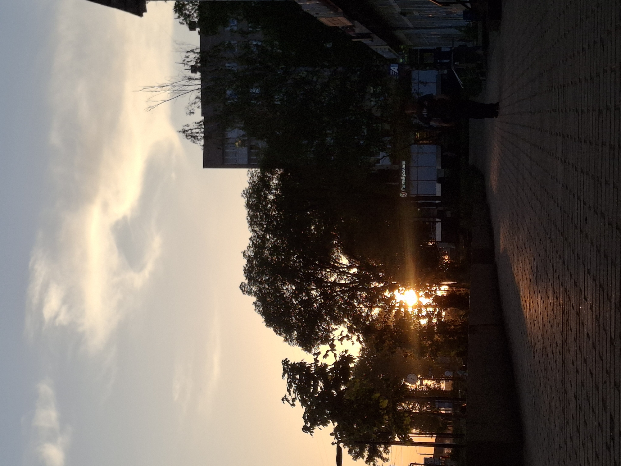 The image shows a street scene at sunset in what appears to be the city of Chaikovsky, Russia. The sky is a pale yellow color with scattered white clouds. The sun is low in the sky, shining through trees and causing bright flares. A building is visible in the background. The sidewalk is made of rectangular paving stones and has a dark-colored edging along the edge. People walk along the sidewalk. Street signs are visible. The sign on the building says "Пятёрочка."