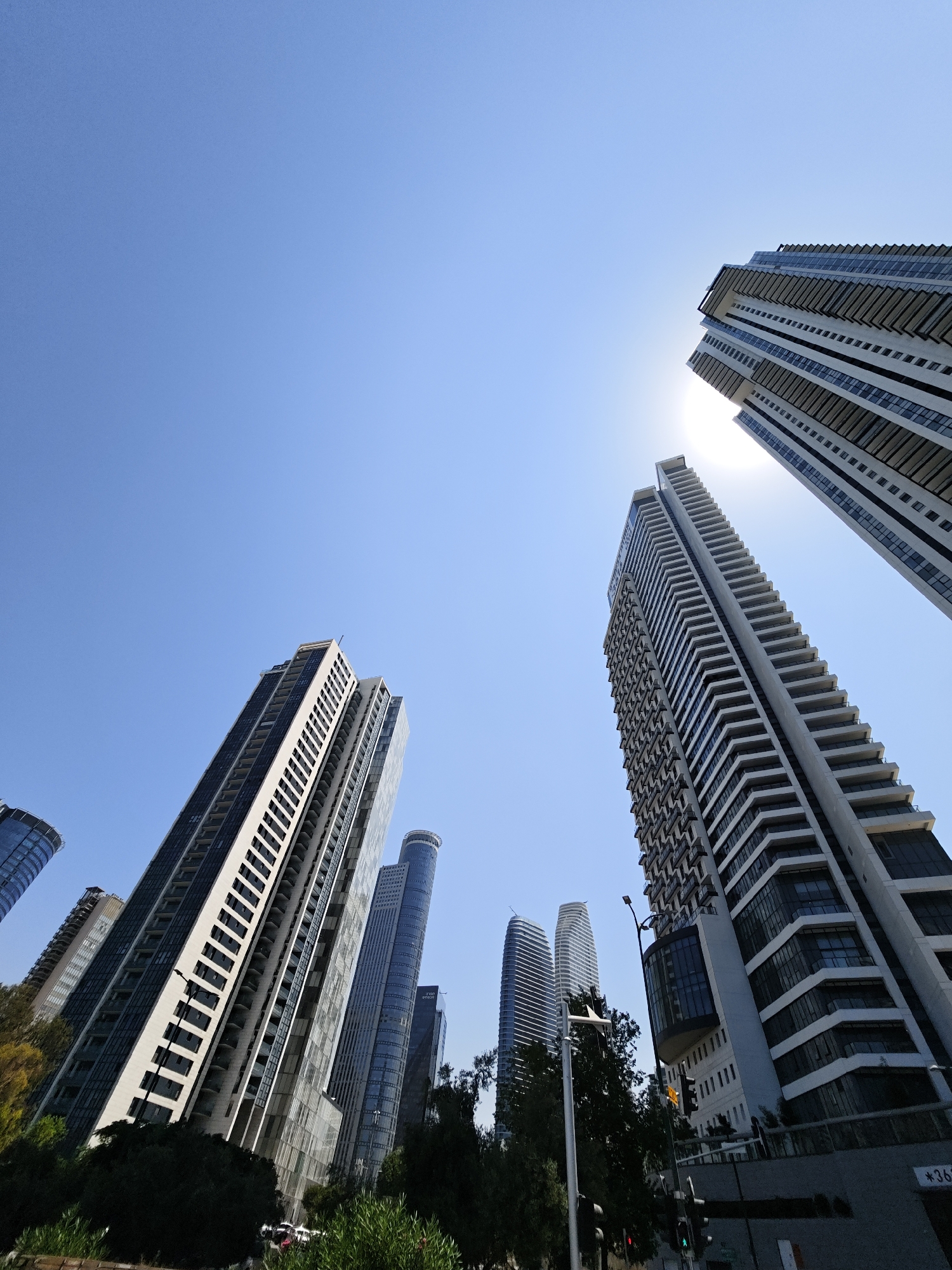 The image shows a low-angle view of modern skyscrapers in Givatayim, Israel, under a clear blue sky. The sun is partially visible, creating a lens flare effect. The buildings are tall and contemporary in design, with many windows and balconies. Some of the buildings have distinctive architectural features, such as curved facades and stepped upper levels. Green trees and foliage are visible at the base of some of the buildings, suggesting a park or landscaped area. The overall impression is of a modern urban environment with a focus on architectural height and design.