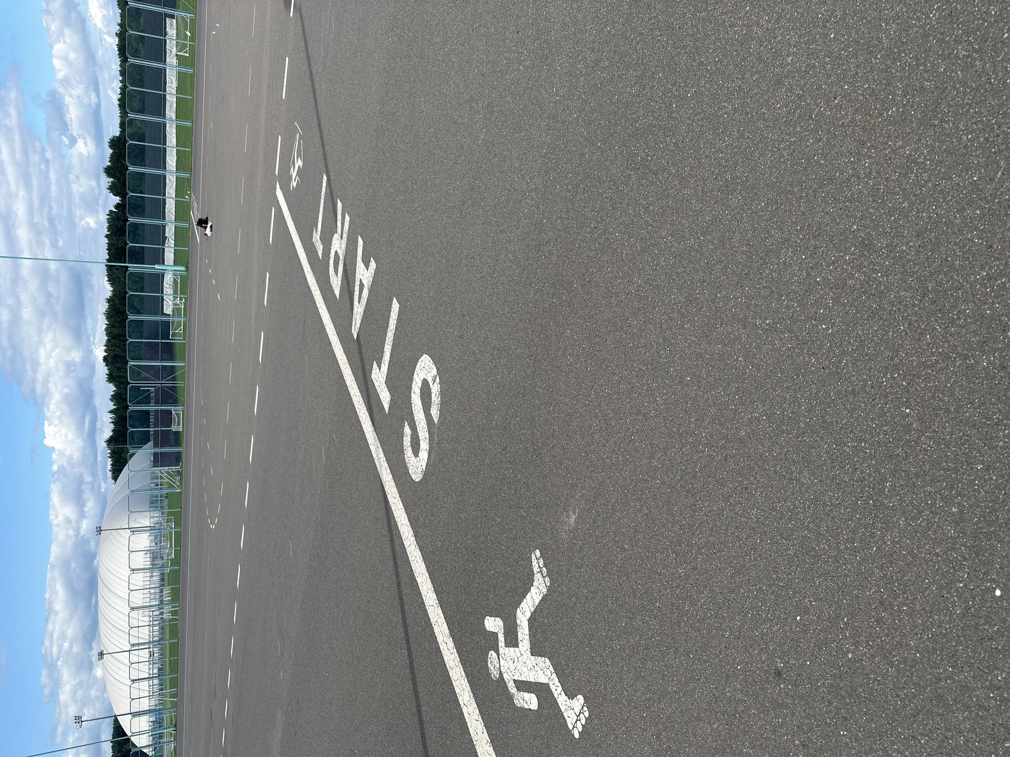 This is a ground-level, wide-angle shot of an outdoor asphalt track or court. The camera is tilted, giving a dynamic perspective. In the foreground, large white painted letters on the asphalt read "START". To the right of the "START" text, a white painted stencil of a rollerblader is visible.
In the mid-ground, a person on roller skates or a scooter is moving along the track, appearing as a small, dark figure. The track is marked with white dashed lines, suggesting lanes or divisions.
In the background, a tall, green metal fence runs horizontally across the image, separating the track from a grassy area. Beyond the fence, a dense line of dark green trees forms the horizon. Above the trees, a bright blue sky is filled with fluffy, white clouds. On the far left of the background, a large, white domed structure, possibly an indoor sports facility or arena, is partially visible. There are also lampposts visible in the distance.
The lighting suggests it is daytime, with clear, bright conditions. The scene appears to be a sports or recreational facility. There is no obvious indication of the time of day or specific weather conditions beyond a generally sunny sky. The overall mood is active and possibly energetic due to the presence of recreational equipment and markings on the track.