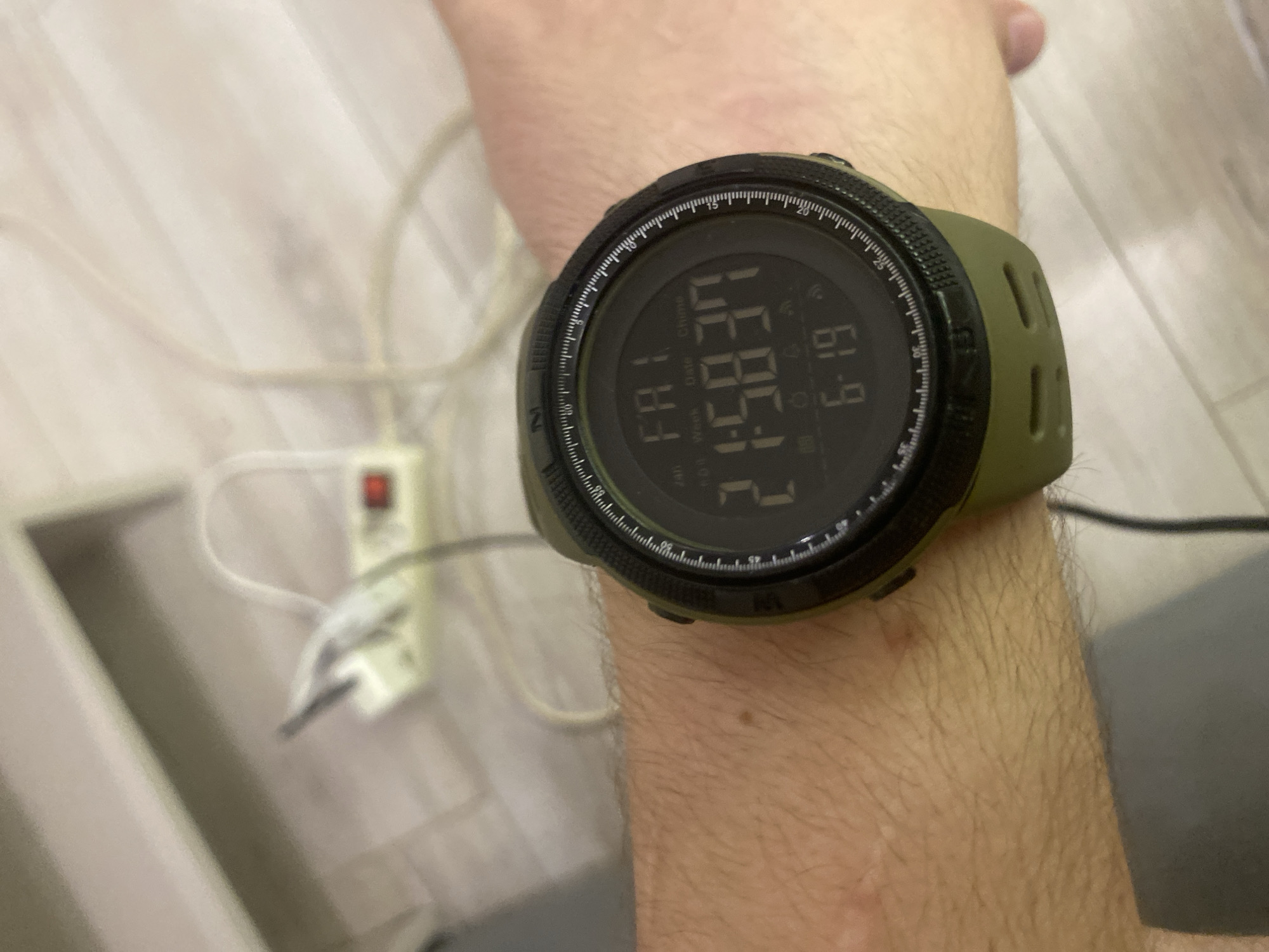 A close-up, eye-level shot shows a digital watch on a person's wrist. The watch is green with a black face and displays the time as 21:58:37 and the date as 6:19. The background is blurred, showing a white power strip with a red switch and electrical cords. The floor appears to be made of light-colored laminate flooring. The image was taken in an indoor setting in Krasnodar, Russia.