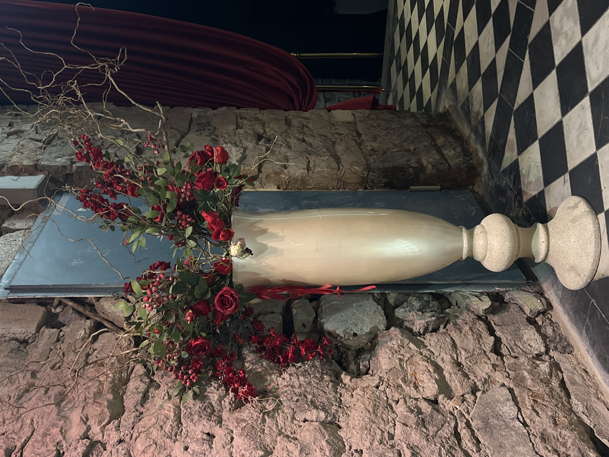 The image depicts a decorative floral arrangement within what appears to be an interior space, possibly a restaurant or event venue in Yerevan, Armenia. The arrangement is placed on a grey ledge made of stacked rough stones, positioned against a wall with a black and white checkered tile pattern. The flowers primarily consist of red roses and small red berries, interspersed with green leaves and bare branches. The flowers are arranged in a tall, cylindrical, off-white vase with a decorative base. Above the stone ledge, draped red fabric is visible. The lighting is somewhat dim, highlighting the floral arrangement. The absence of people indicates the scene is static. The combination of materials (stone, flowers, fabric, tile) suggests a contrasting design aesthetic.